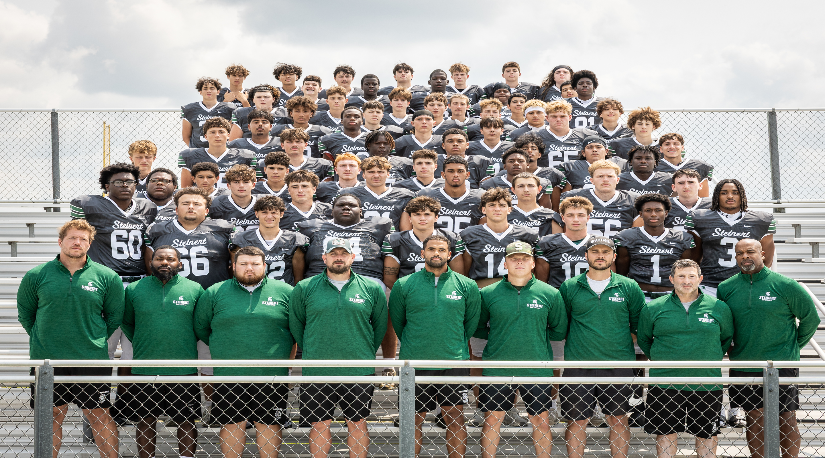 A large group of young men in football uniforms pose for a photo on bleachers in front of a chain-link fence.