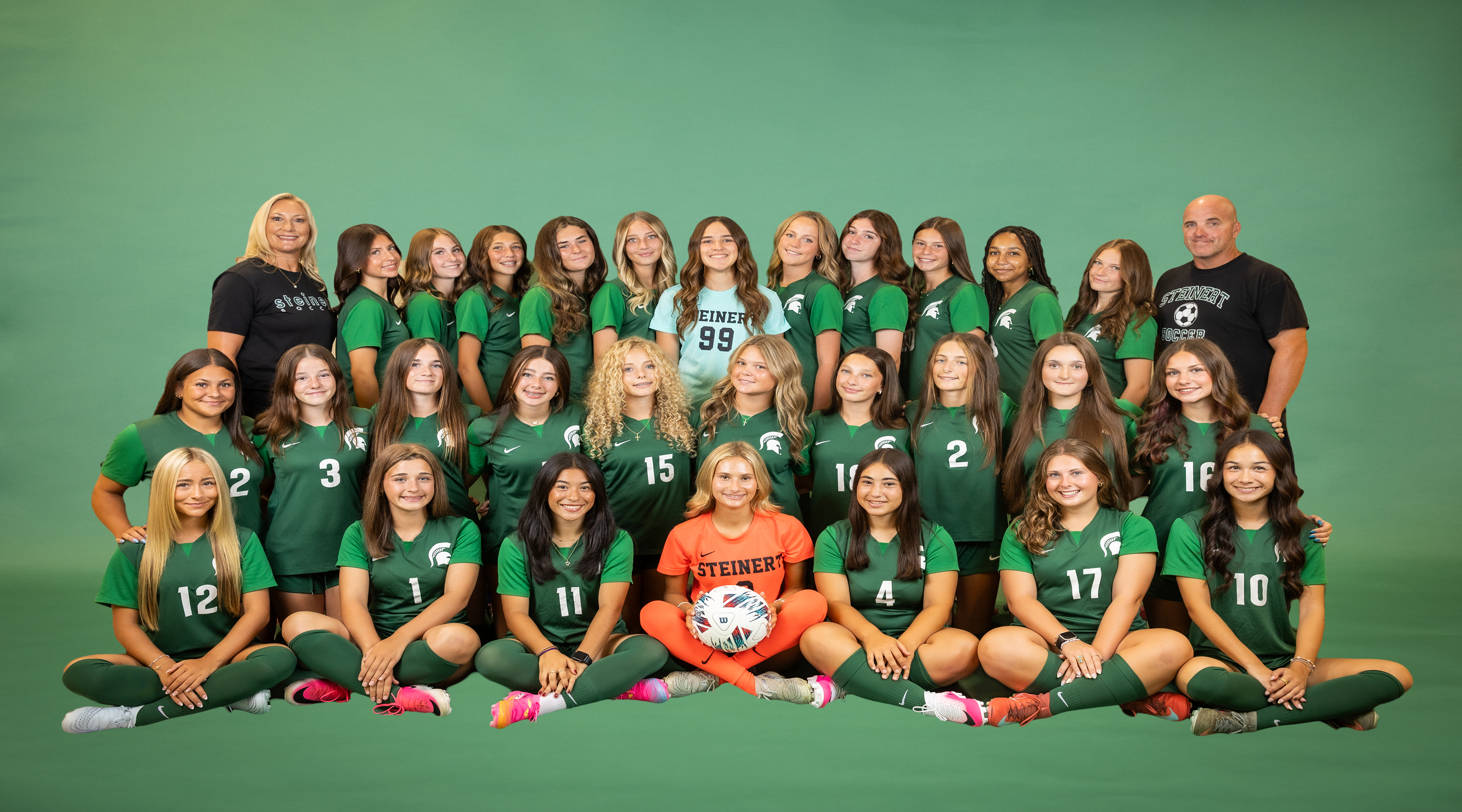 A girls' soccer team poses for a photo in front of a green backdrop, with players in green uniforms in the foreground and coaches standing behind them.