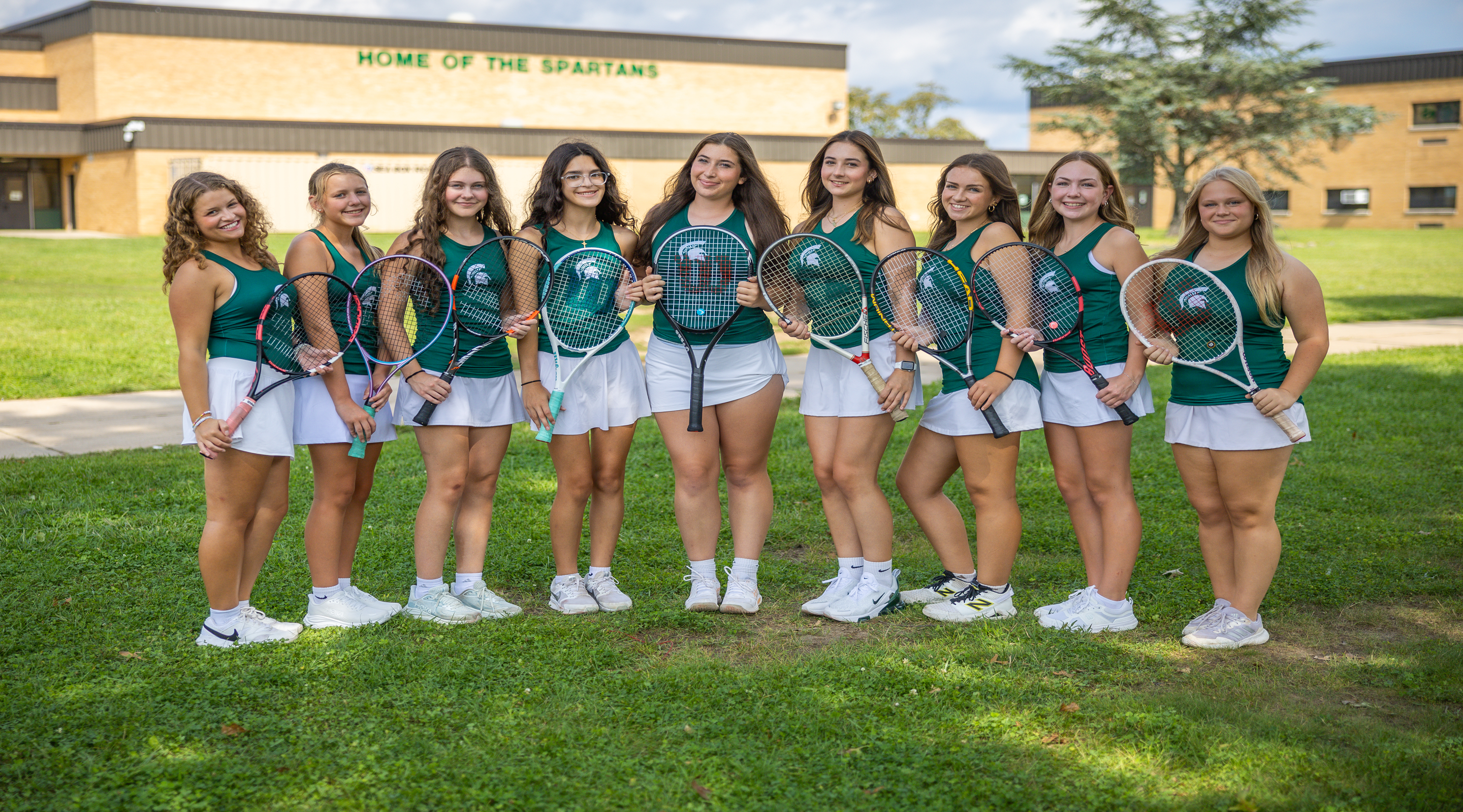A group of eight young women in tennis uniforms stand on a grassy field holding rackets, with a school building labeled "HOME OF THE SPARTANS" in the background.
