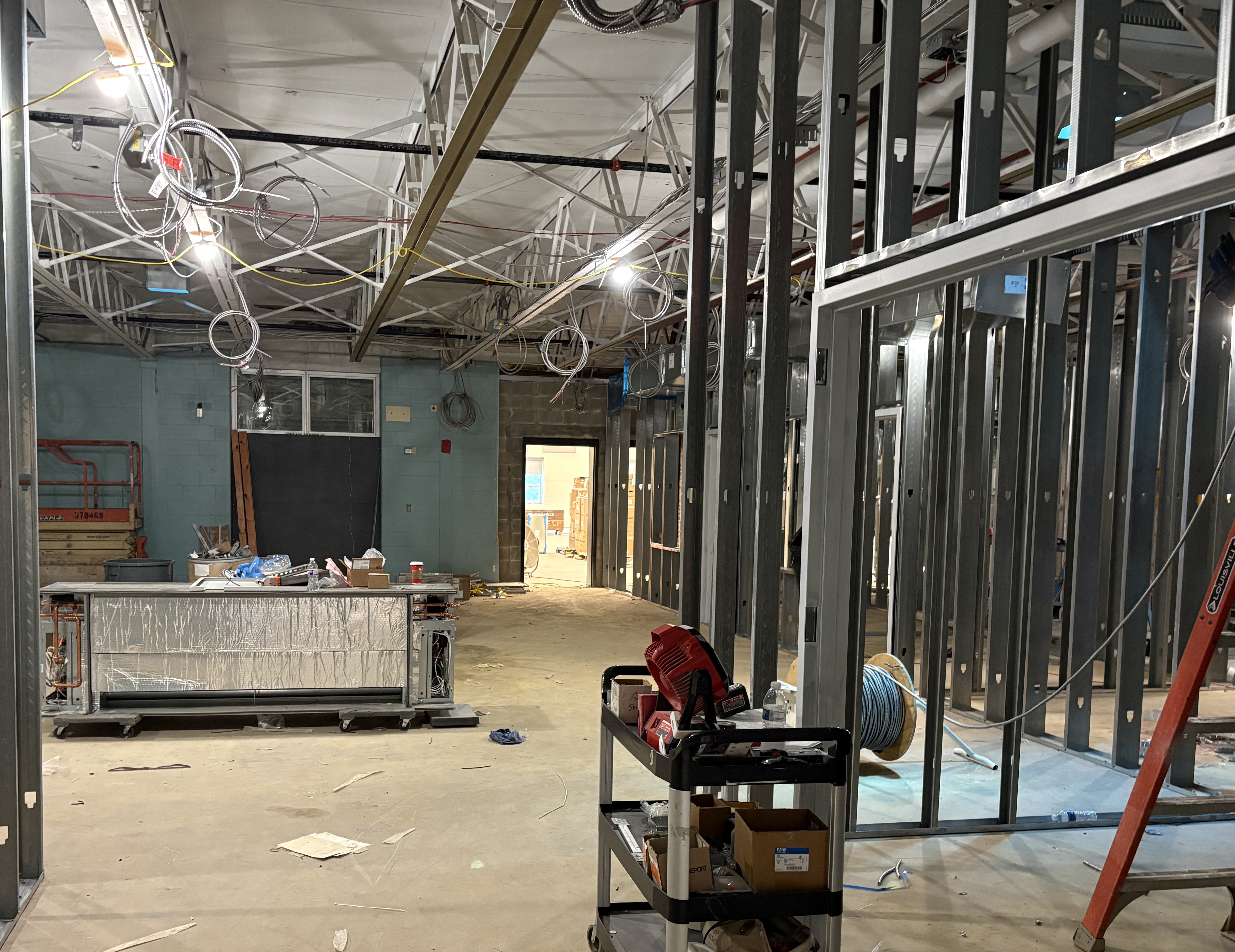 A construction site with metal studs and exposed wiring in the foreground, and a hallway with doorways leading to other rooms in the background.