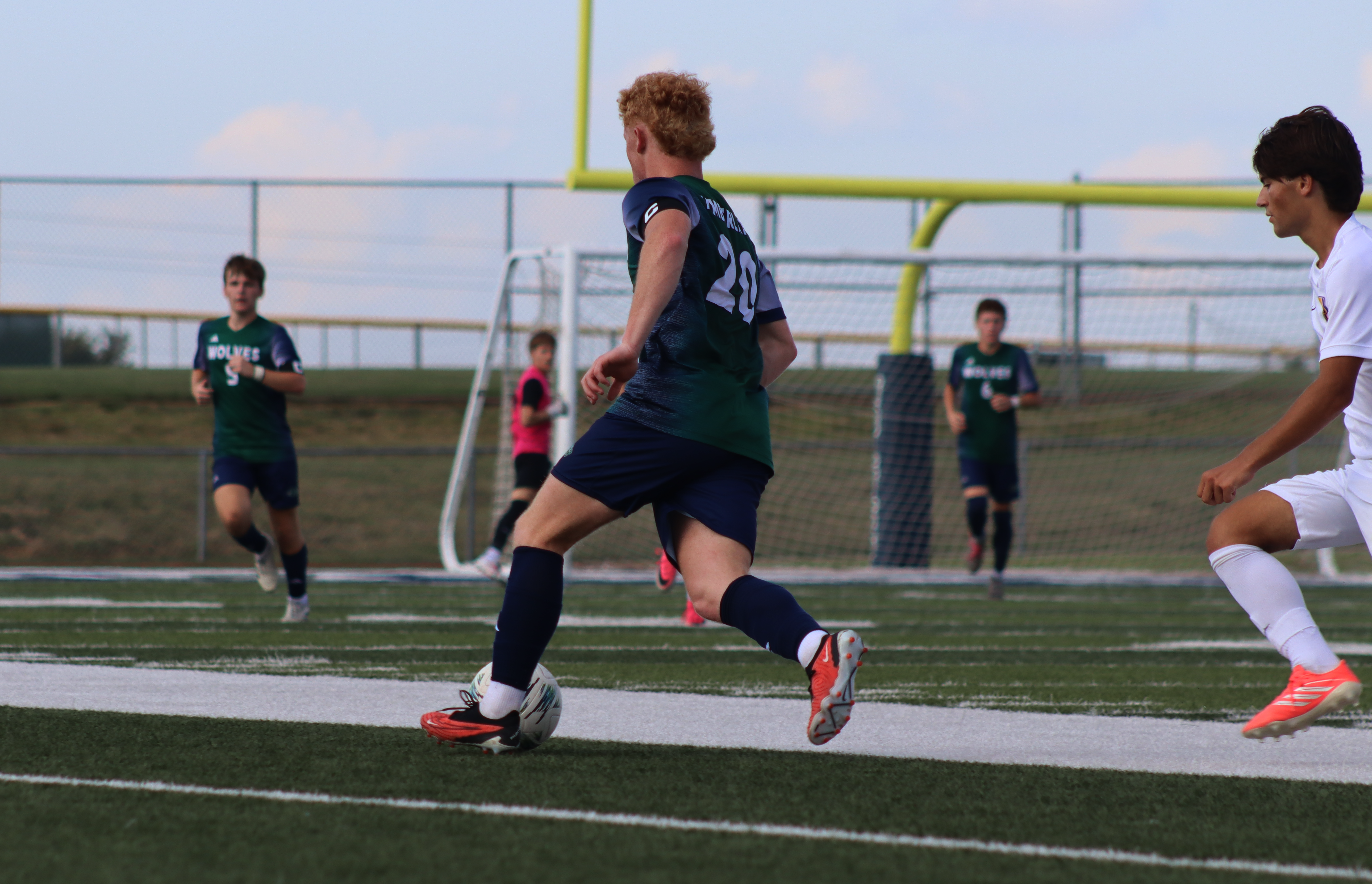 A soccer player in a green jersey dribbles a ball in the foreground while other players and a goal stand on a grassy field under a clear sky.