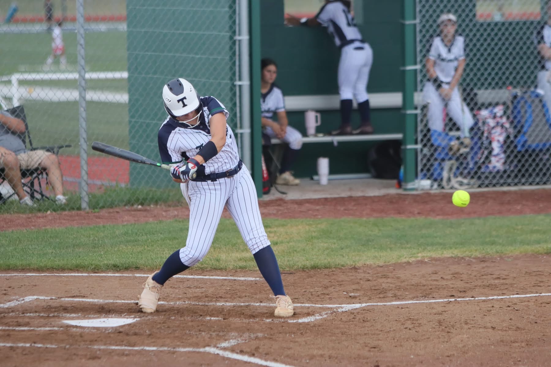 A softball player in a pinstriped uniform swings a bat at a yellow ball in the foreground, with teammates and a dugout visible in the background.
