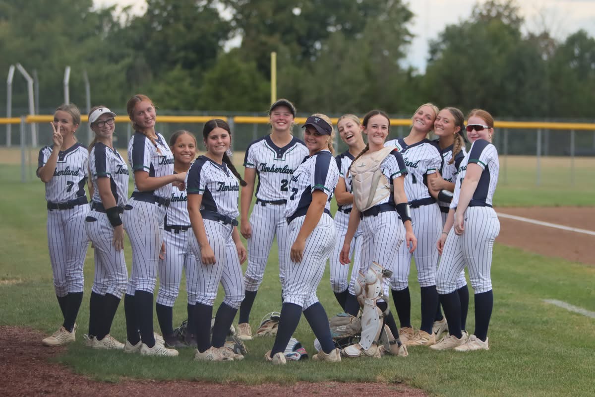 A group of female softball players in pinstriped uniforms pose together on a grassy field with a line of trees in the background.