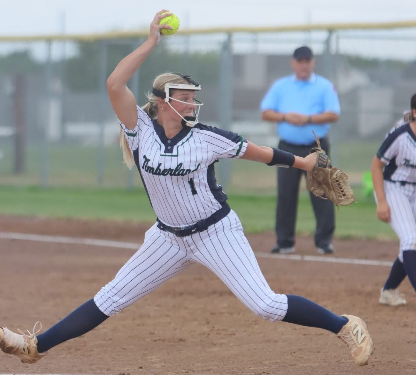 A softball pitcher in a pinstriped uniform winds up to throw a yellow ball on a dirt field with an umpire and another player in the background.