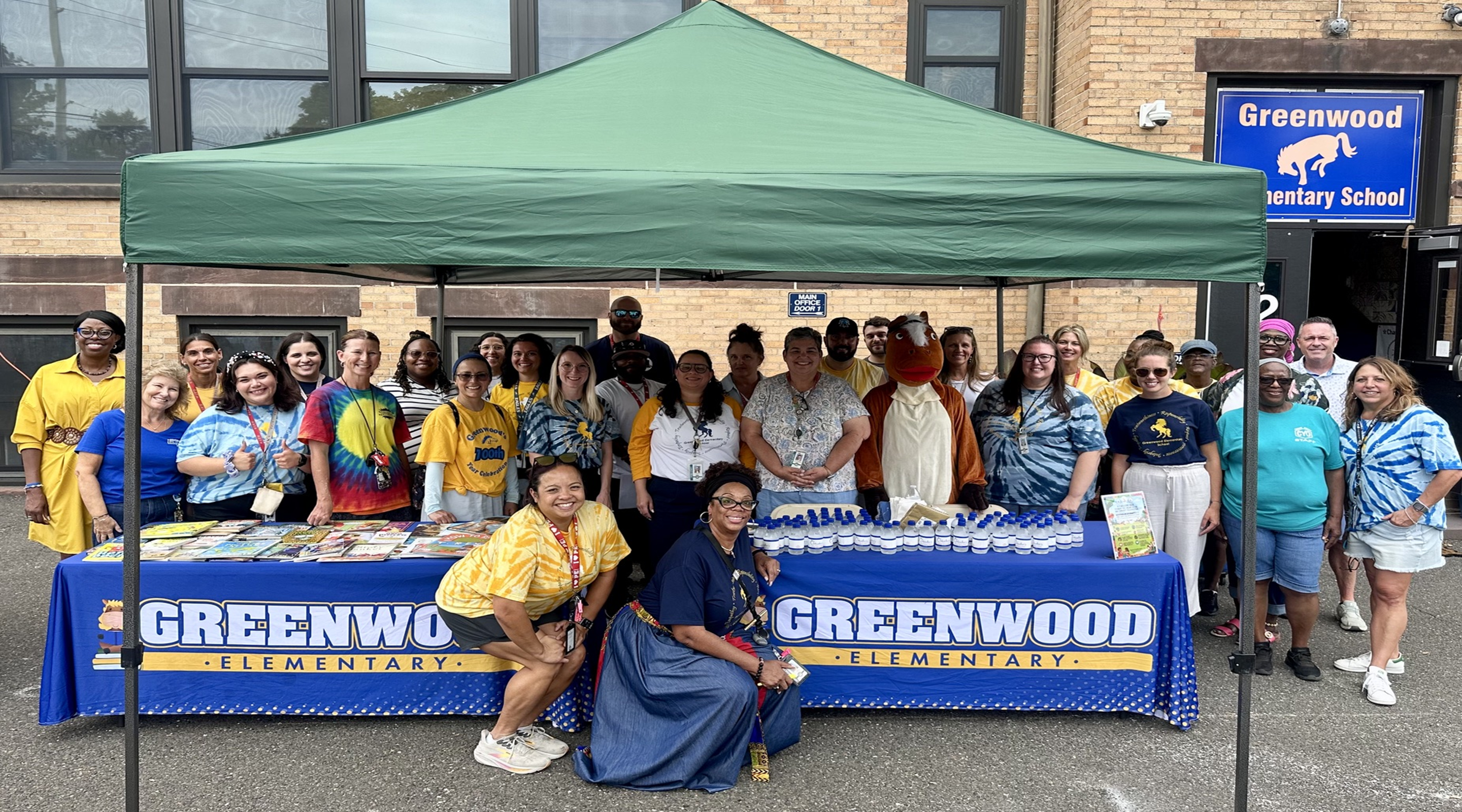 A group of people and a mascot stand behind a blue table under a green canopy in front of Greenwood Elementary School.