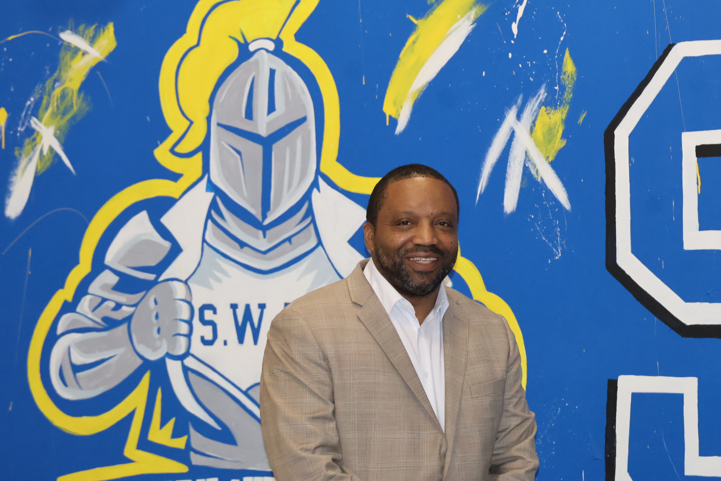 A smiling man in a tan blazer stands in the foreground against a blue background featuring a large graphic of a knight.