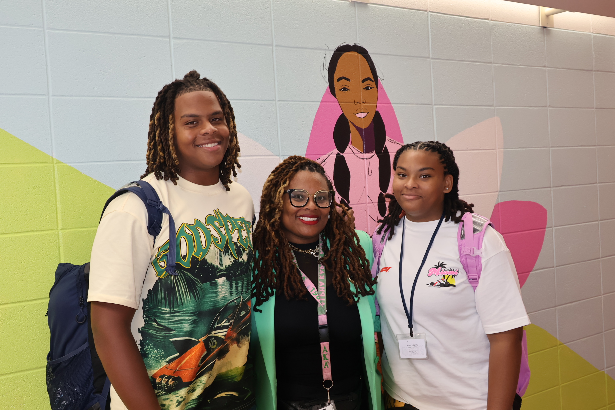 Three people smile for a photo in the foreground against a white tiled wall featuring a colorful mural in the background.