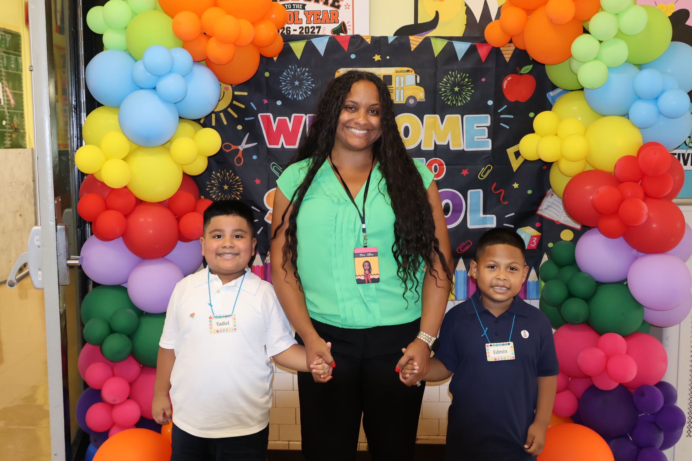 A woman stands between two young boys in the foreground, posed in front of a "Welcome to School" banner and colorful balloon arches in the background.