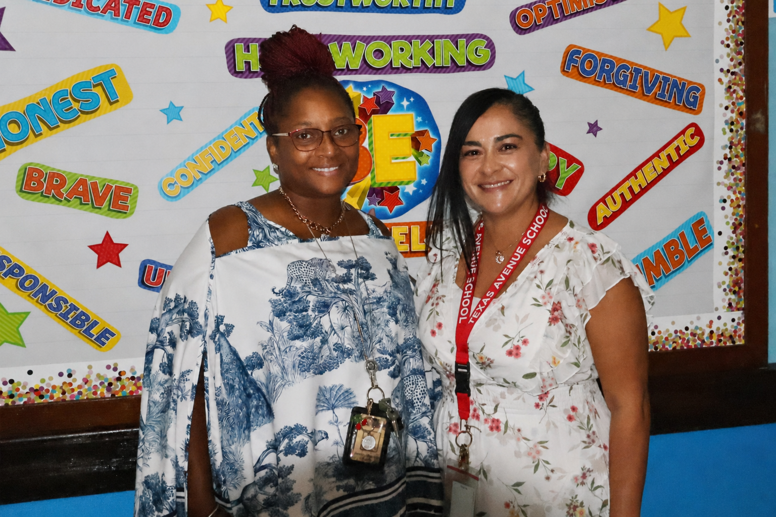 Two women stand smiling in the foreground against a white wall decorated with colorful positive trait words and stars in the background.