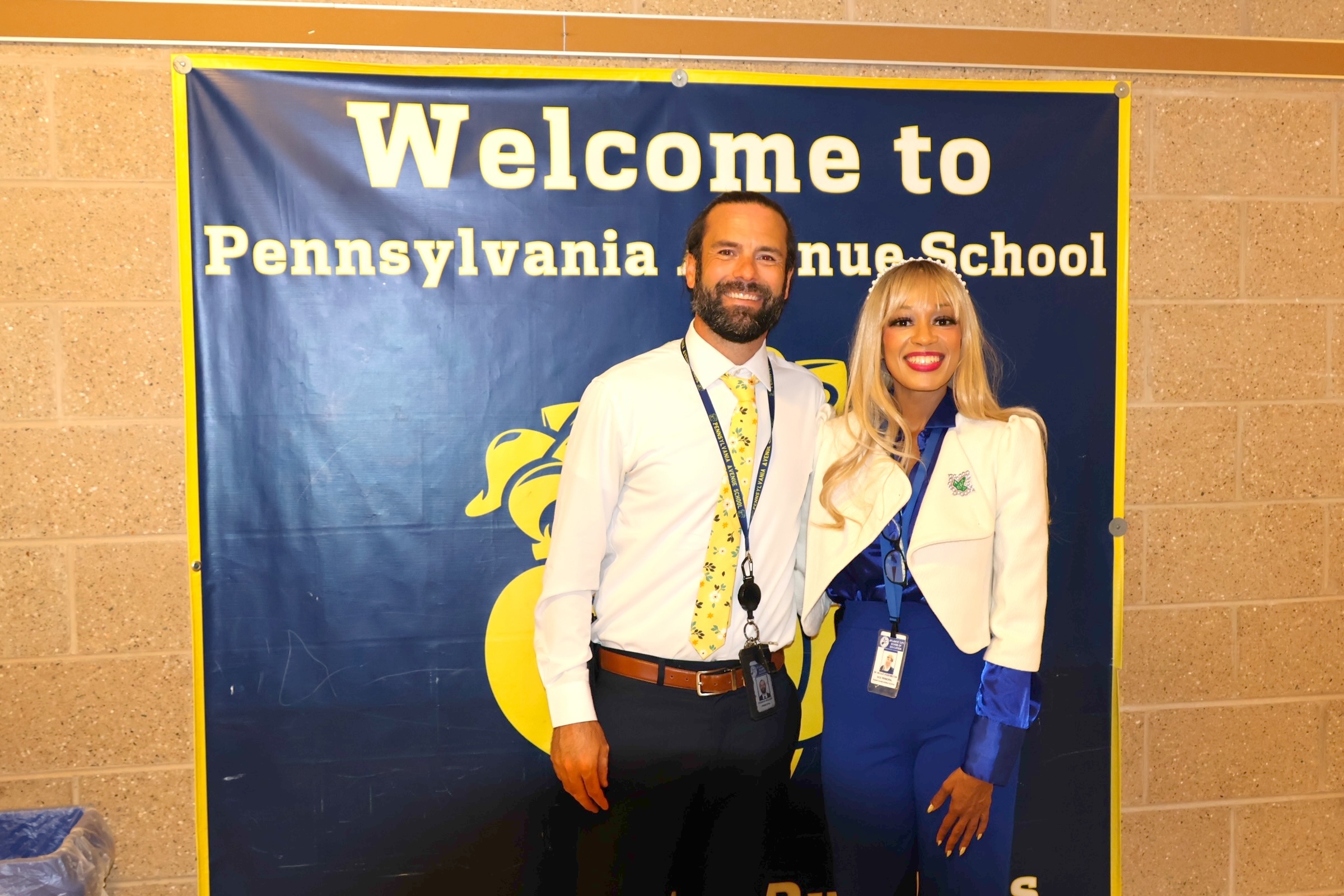 A man in a white shirt and tie and a woman in a white blazer and blue pants stand in front of a blue "Welcome to Pennsylvania Avenue School" banner.