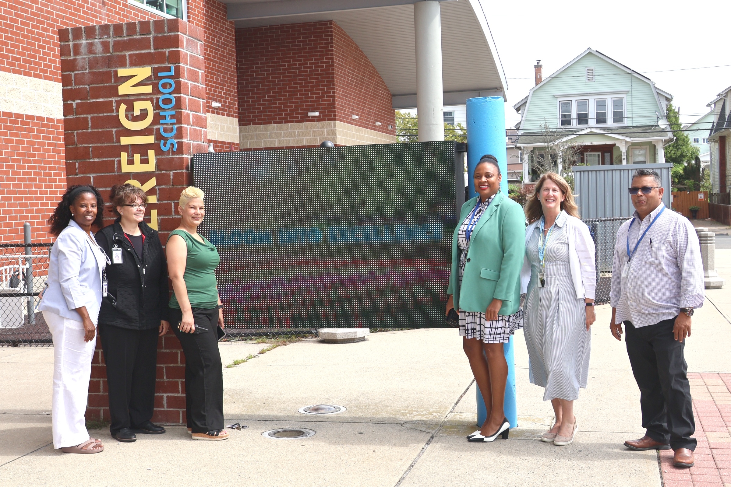 Six people stand in the foreground on a sidewalk in front of a brick building and a large floral mural.