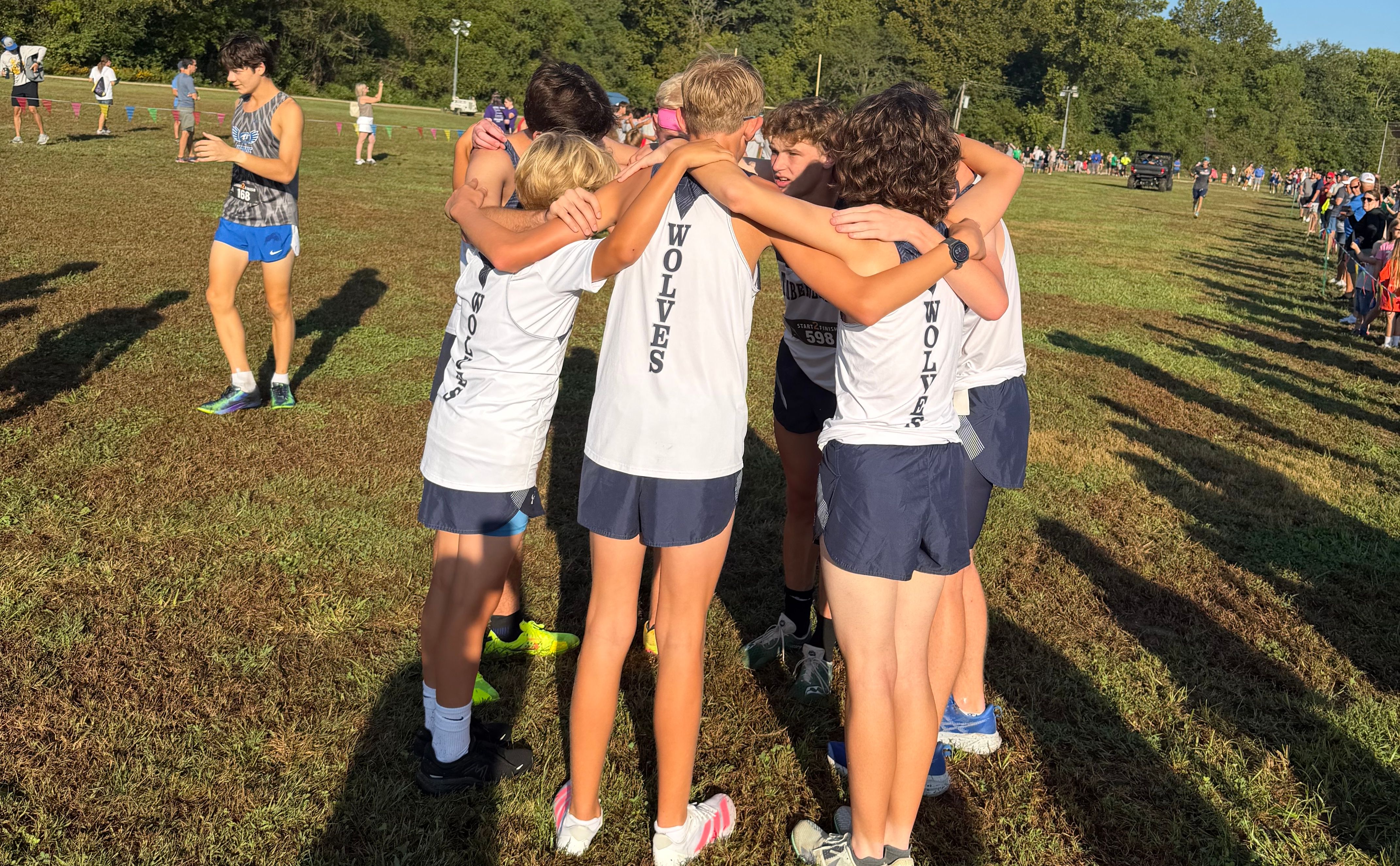 A group of runners in white and blue uniforms huddle together in a grassy field with other athletes and trees in the background.