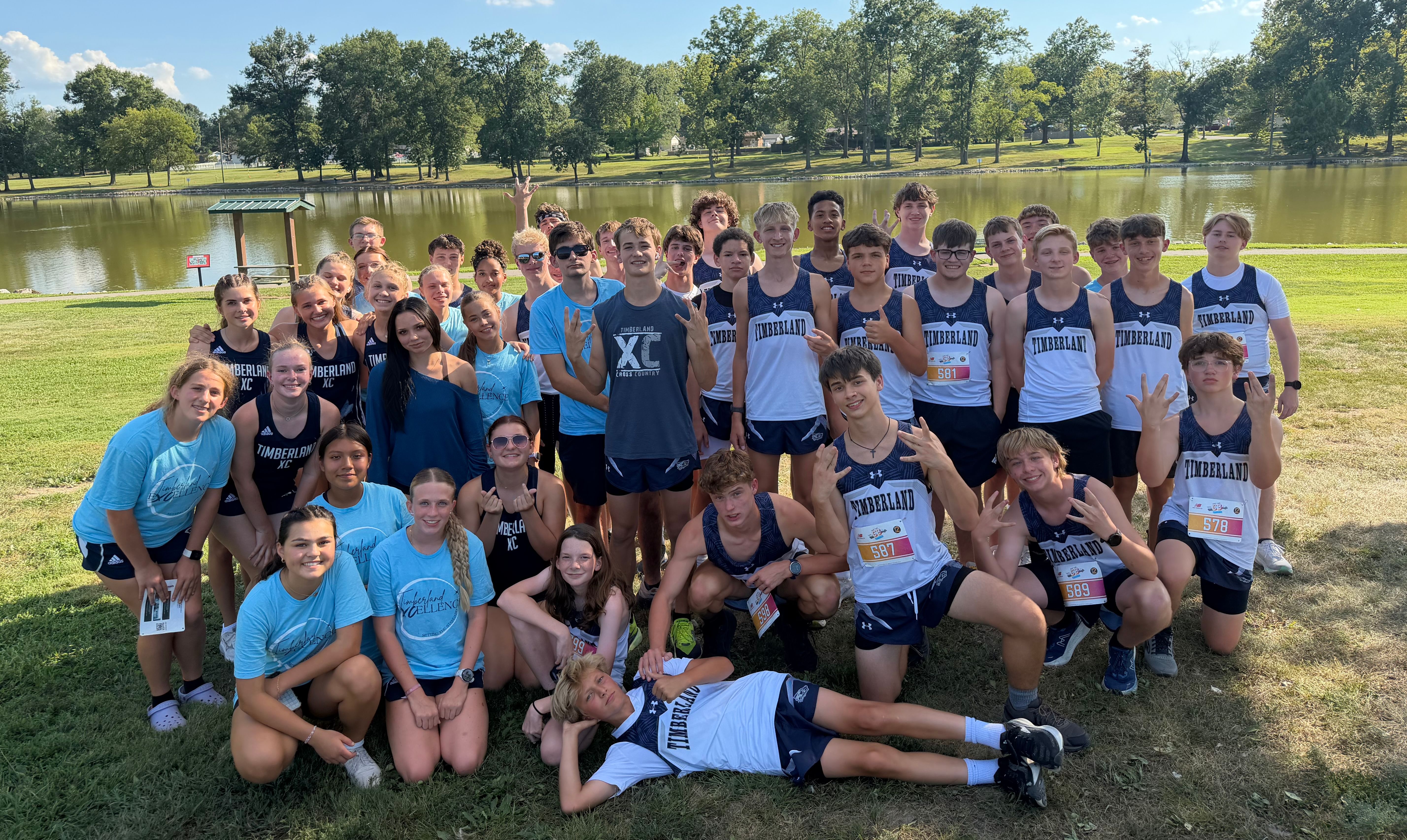 A large group of athletes in blue and white uniforms poses for a photo on a grassy field with a calm lake and lush green trees in the background.