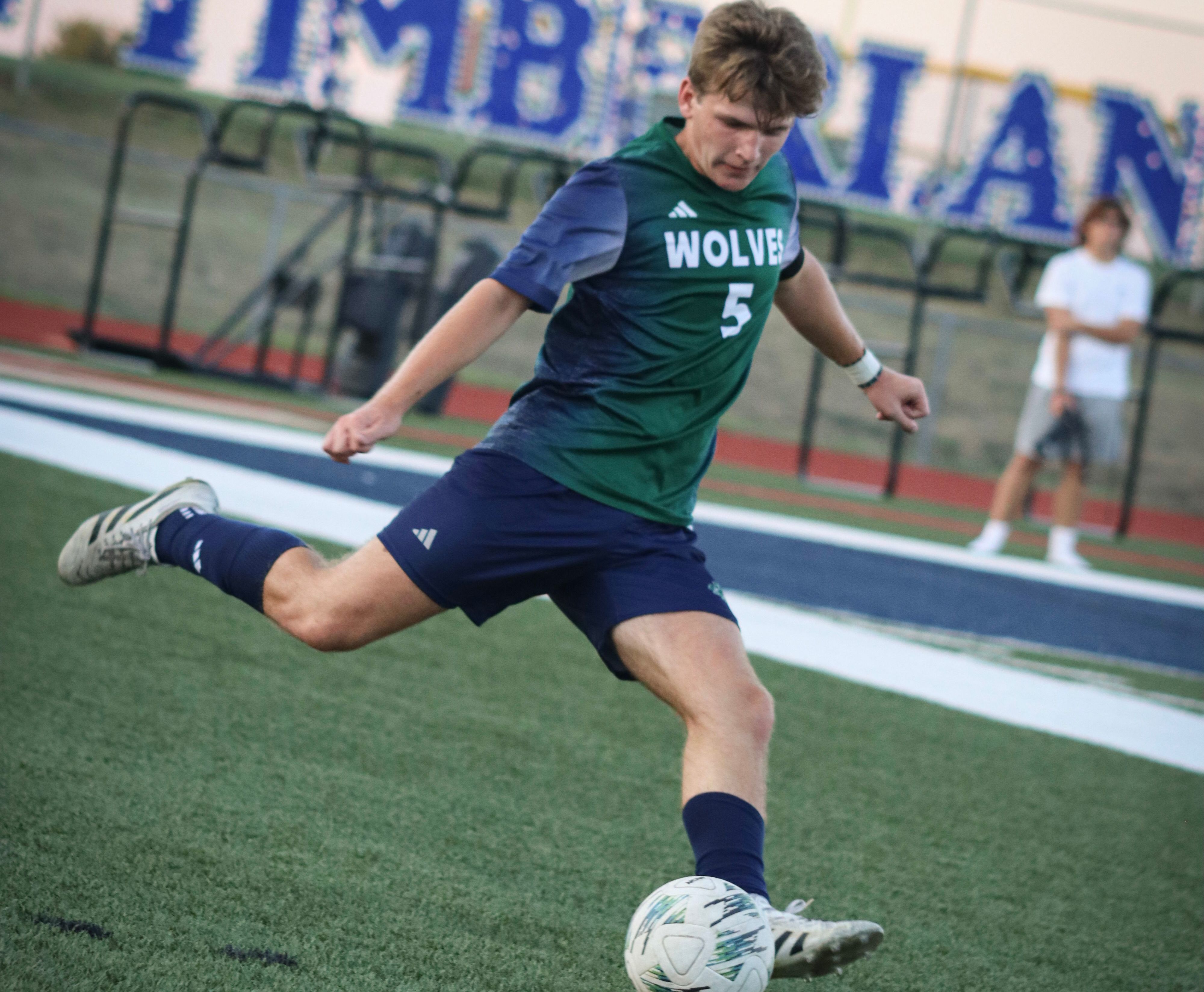 A soccer player in a green and blue uniform kicks a ball on a turf field with a spectator and a large sign in the background.