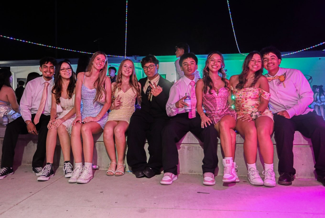 A group of dressed-up teenagers sit together on a concrete ledge against a dark night sky accented by string lights.