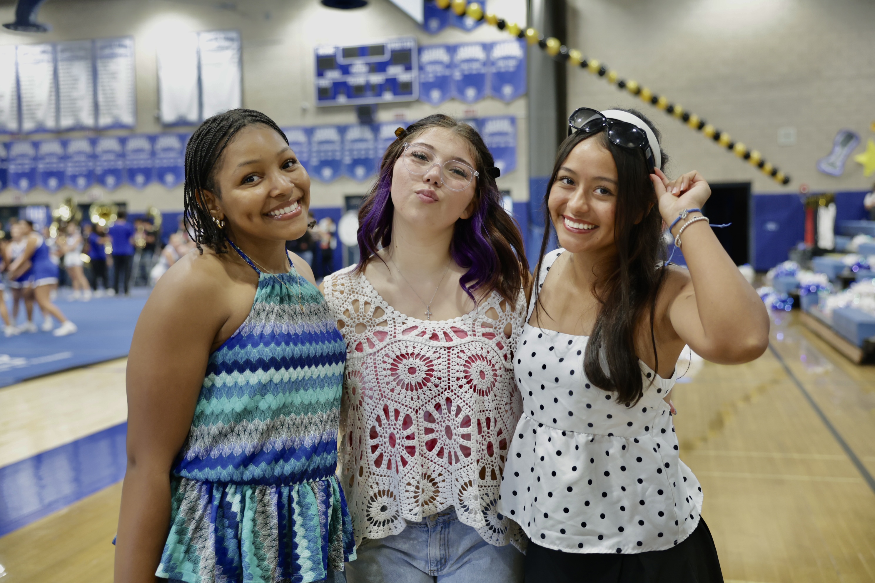 Three smiling young women stand together in the foreground of a gymnasium decorated with blue banners and equipment.