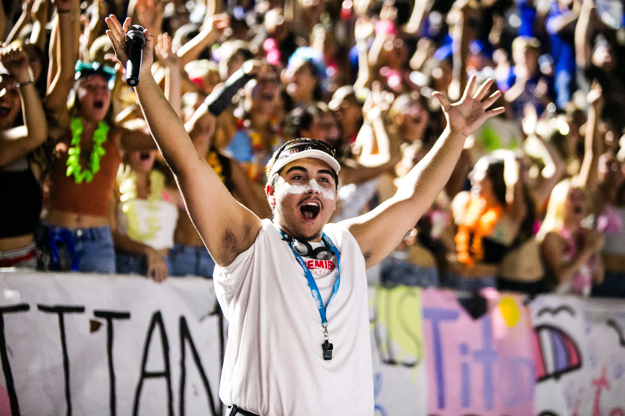 A man with face paint and a white tank top cheers with arms raised in front of a crowded, energetic audience.