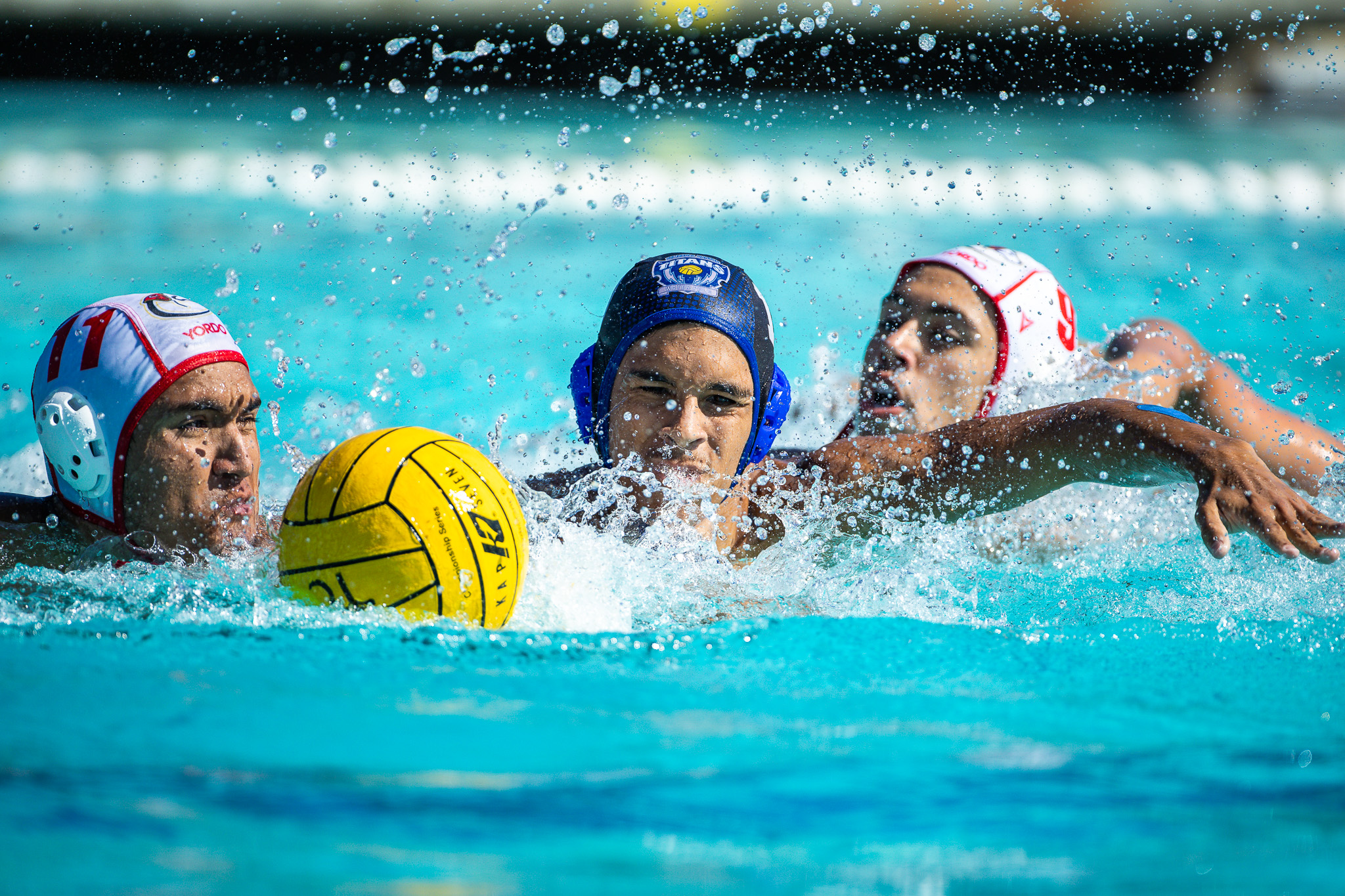 Three water polo players compete for a yellow ball in a splashing blue pool.