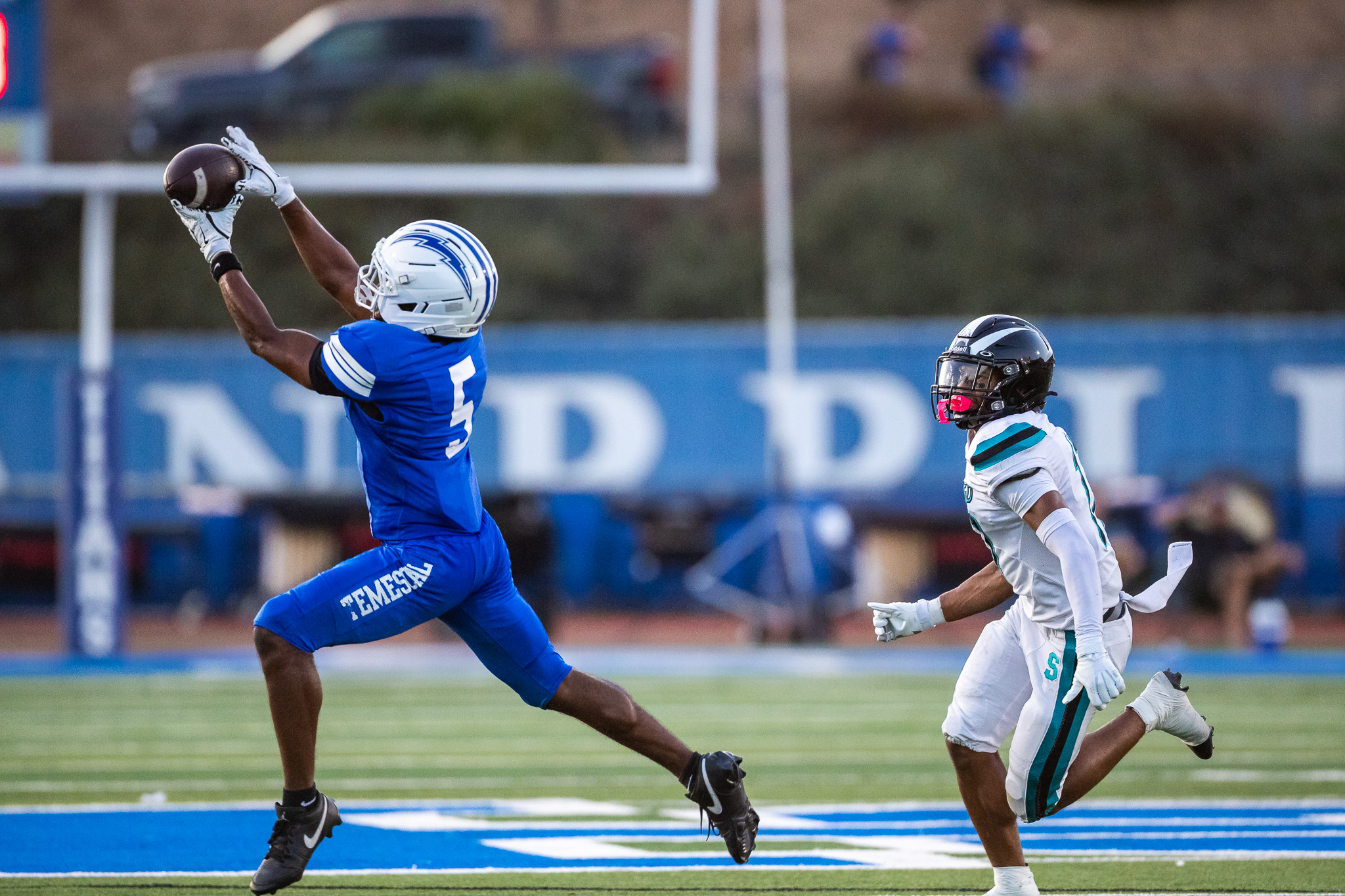 A football player in blue catches a ball while being pursued by a player in white on a green field with a blurred stadium background.