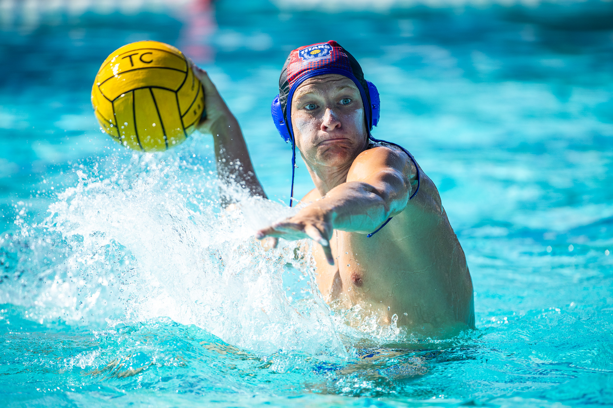 A water polo player wearing a blue and red cap throws a yellow ball amidst splashing water in a bright blue swimming pool.