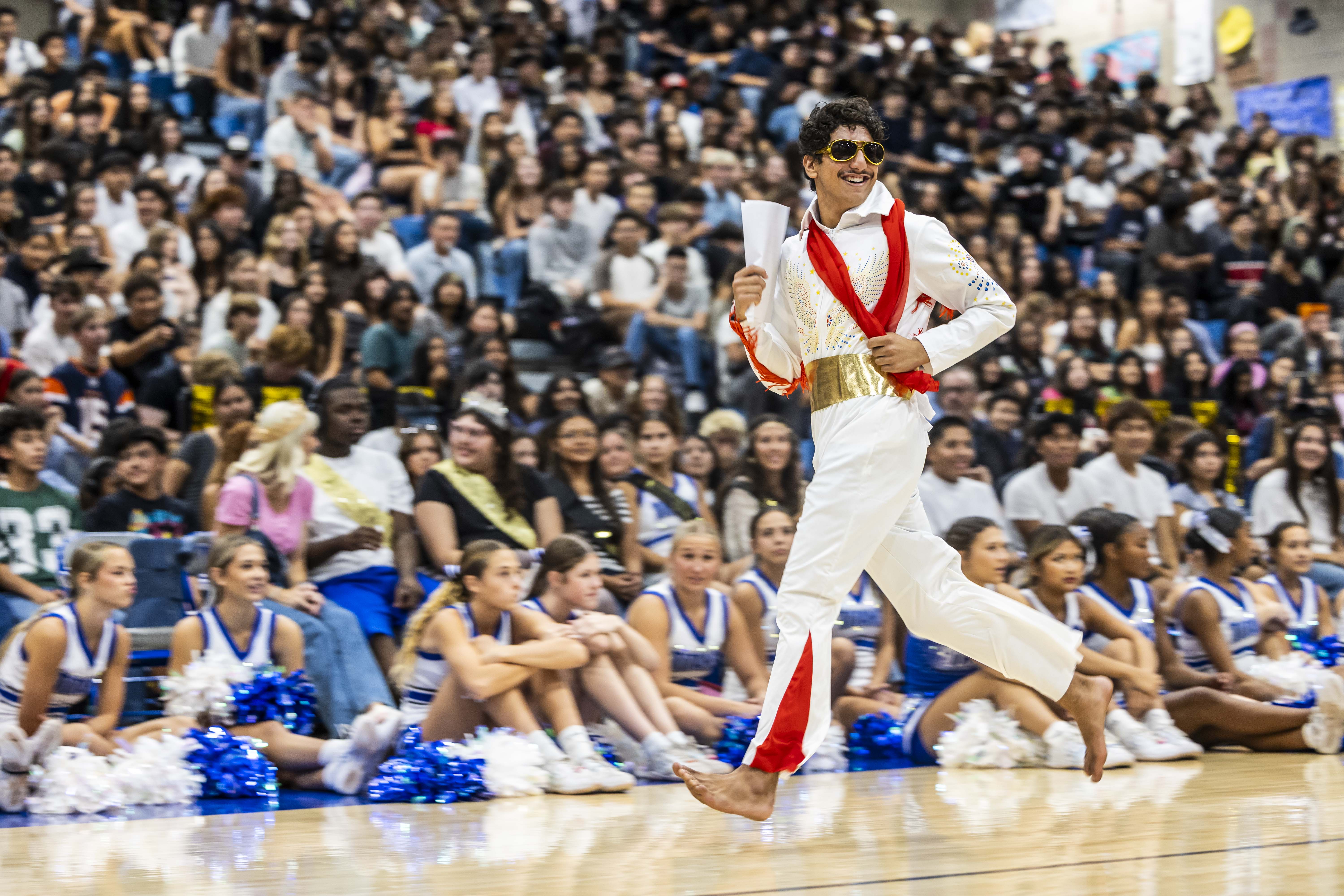 A man in a white outfit with red accents and sunglasses dances in the foreground before a large crowd of students and cheerleaders in a gymnasium.