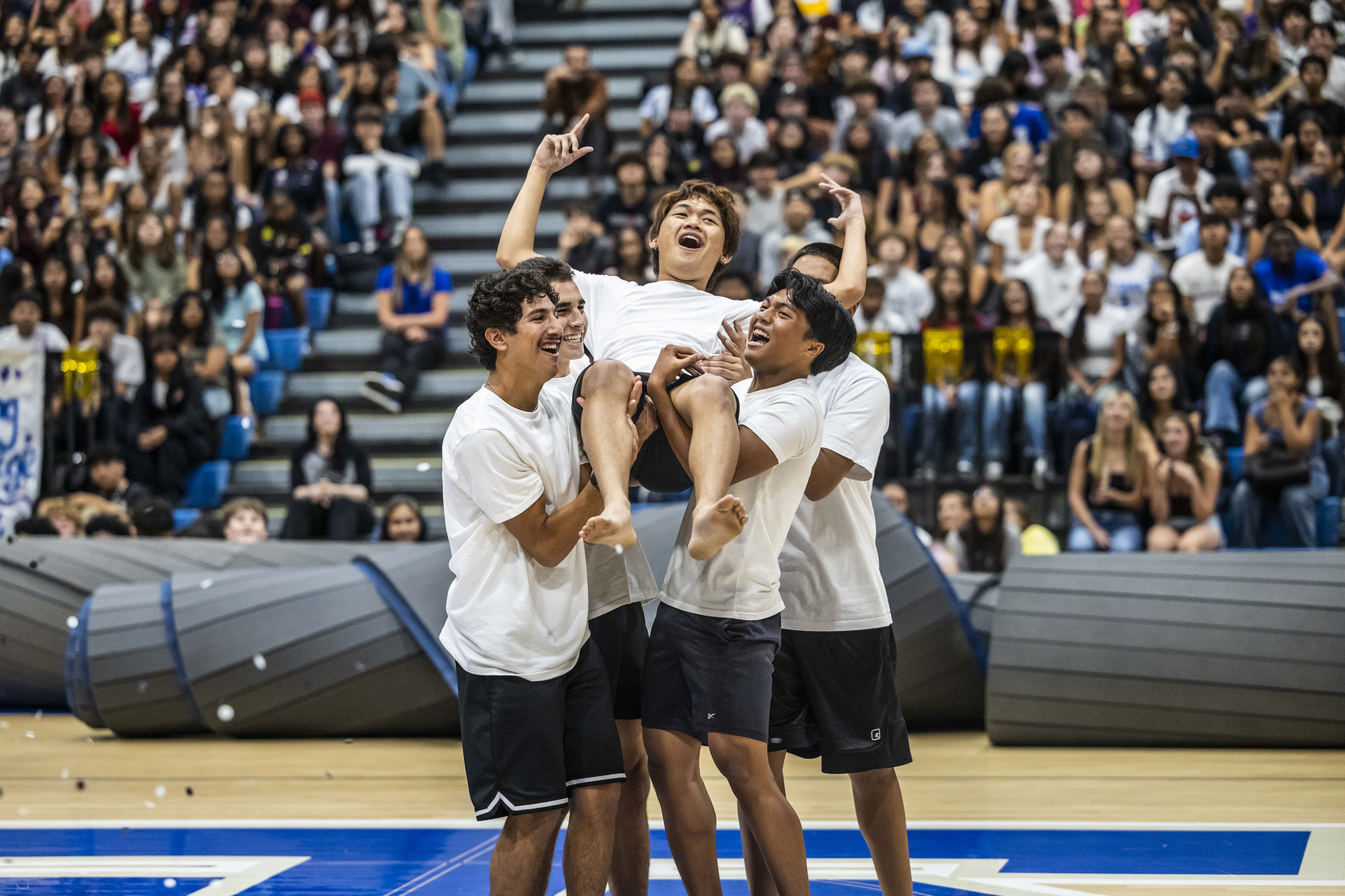 Three young men in white shirts and dark shorts lift a teammate in celebration on a basketball court before a crowded stadium of spectators.