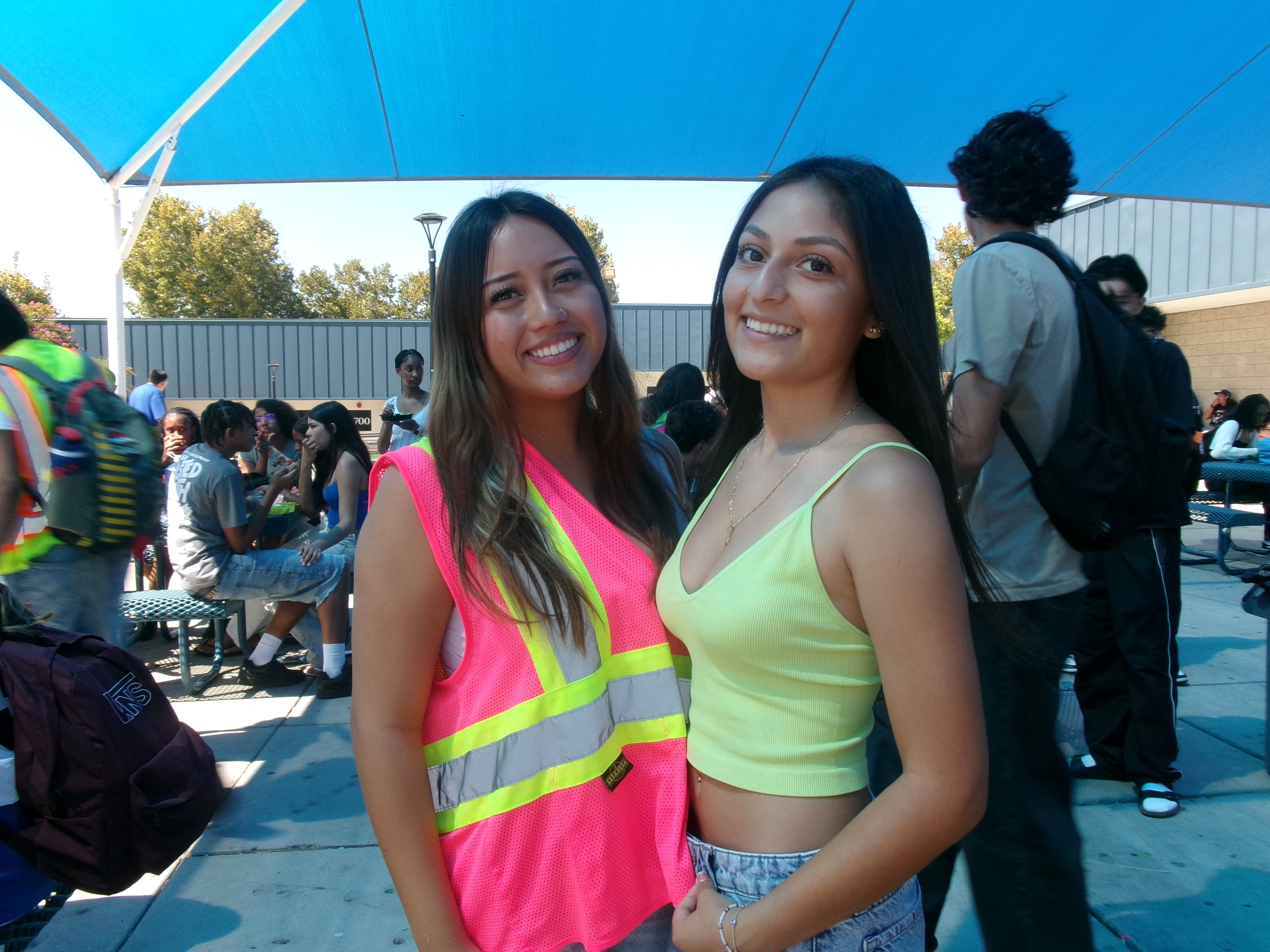 Two smiling young women stand in the foreground under a blue canopy, with other students and backpacks visible in the background.