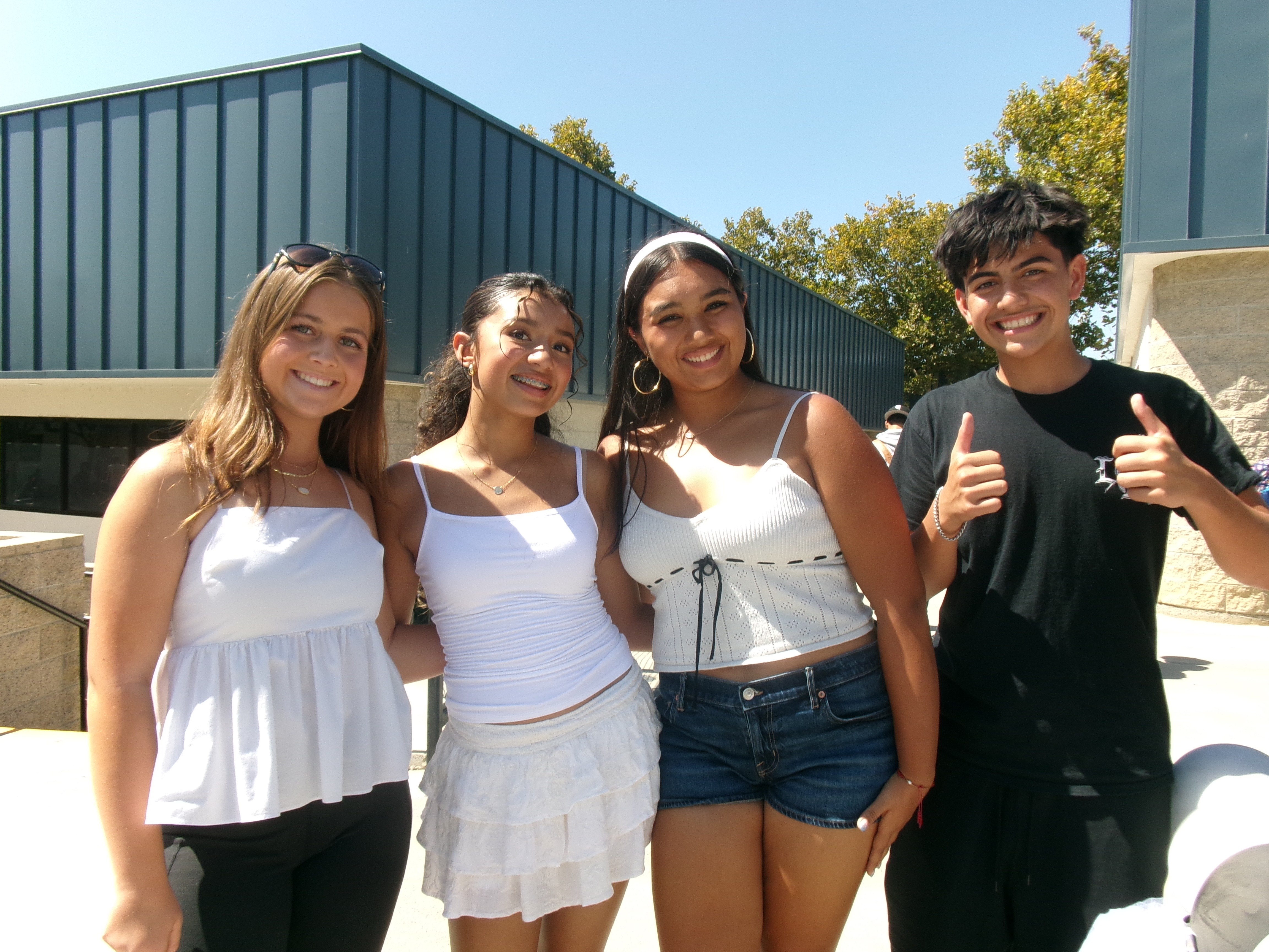 Four smiling young people pose together in the foreground against a background of a modern building and green trees under a clear blue sky.