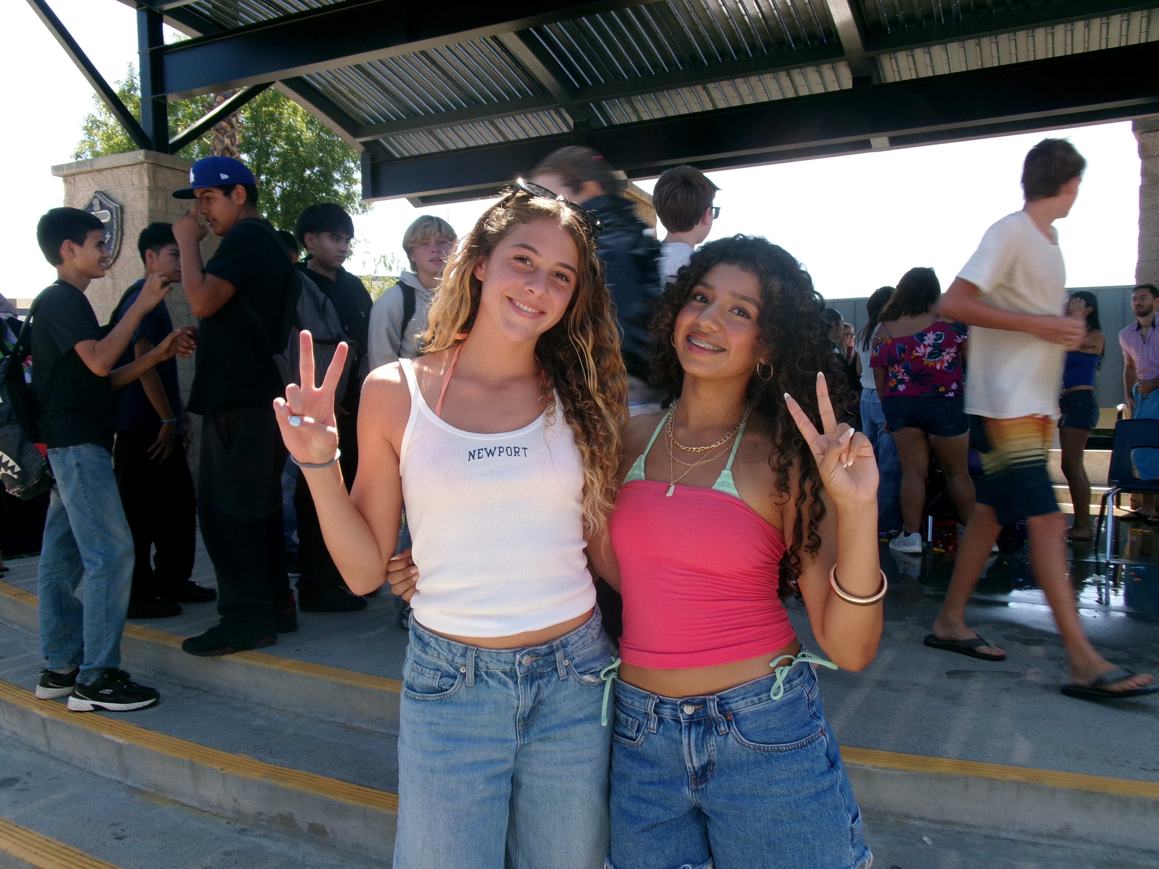 Two smiling young women make peace signs in the foreground while other teenagers stand and walk under a covered outdoor area in the background.