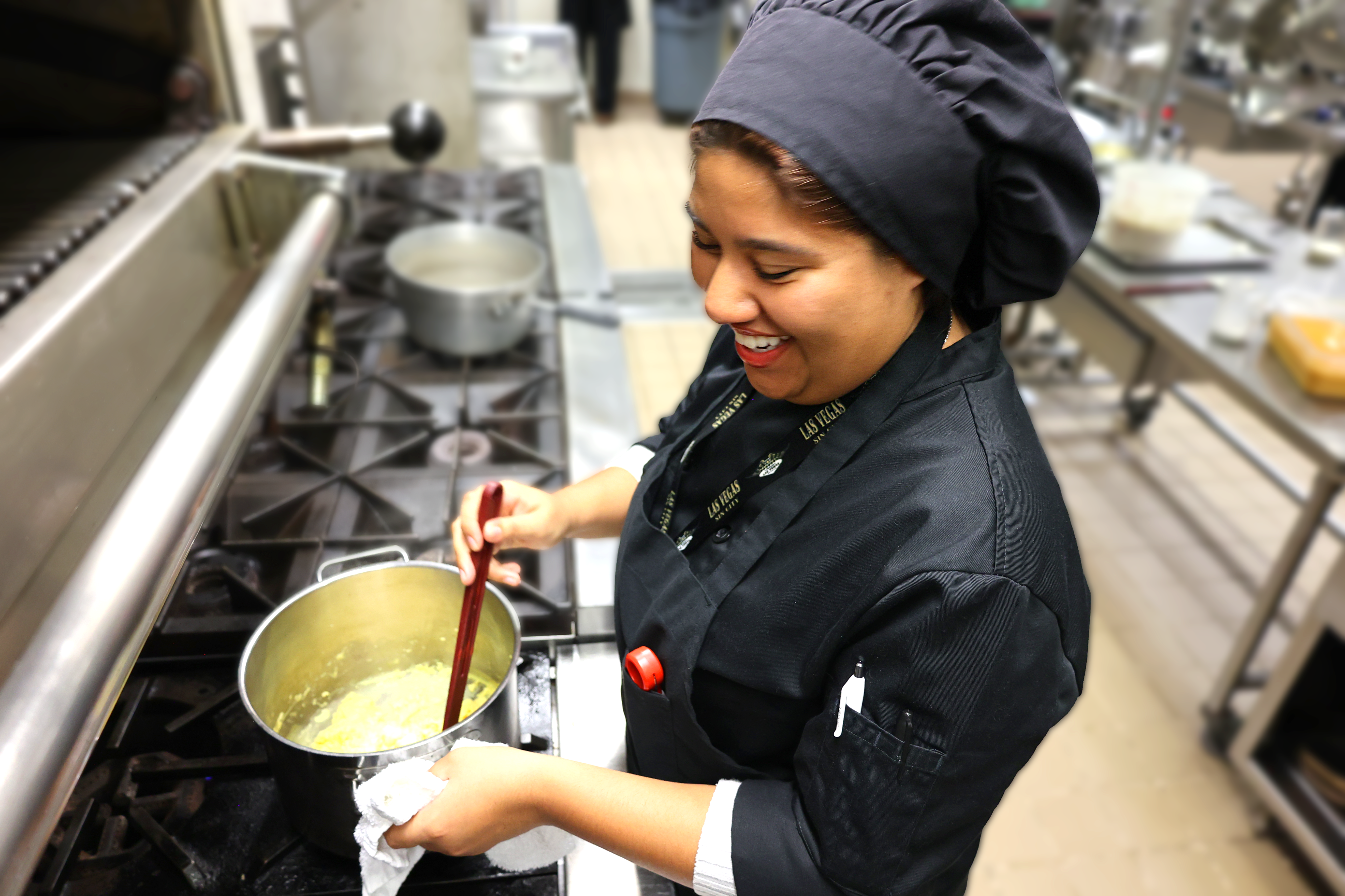 A smiling chef in a black uniform stirs a pot on a commercial stove within a professional kitchen.