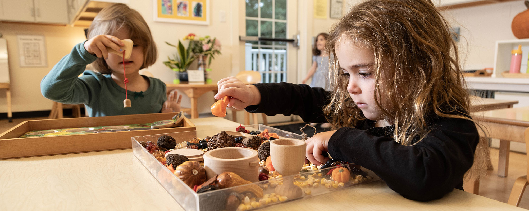 Two young girls play with natural materials in a sensory bin in the foreground of a bright, airy classroom.