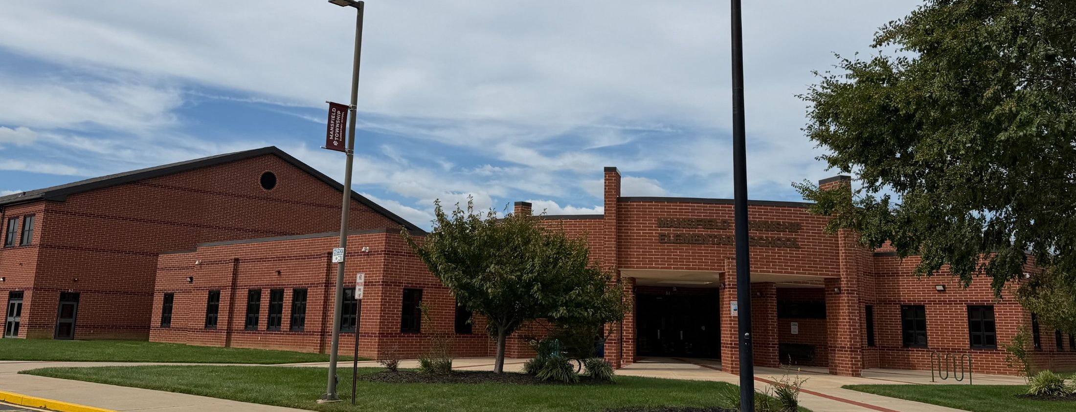 A red brick building stands behind a green lawn and a few trees under a cloudy blue sky.
