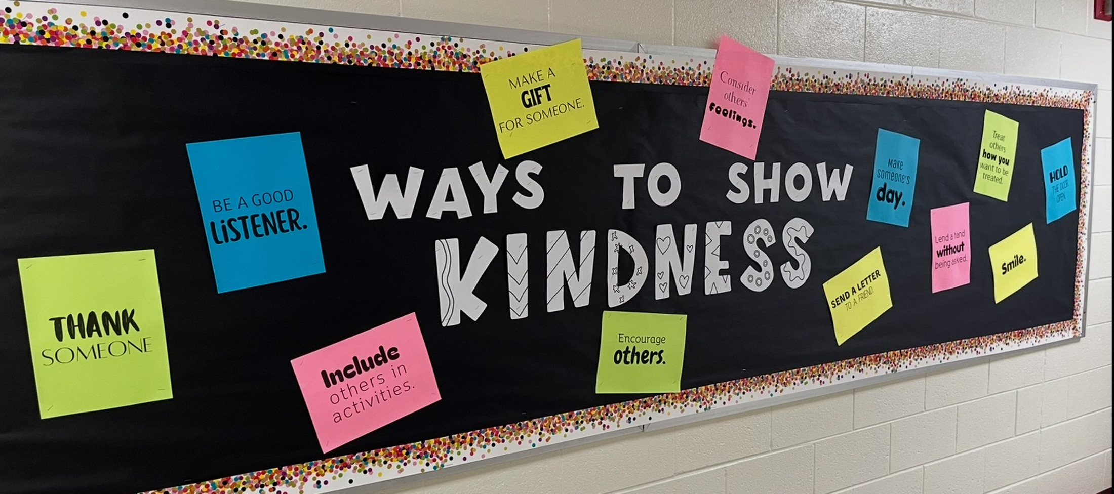 A black bulletin board with the text "WAYS TO SHOW KINDNESS" and various colorful sticky notes is mounted on a white brick wall.