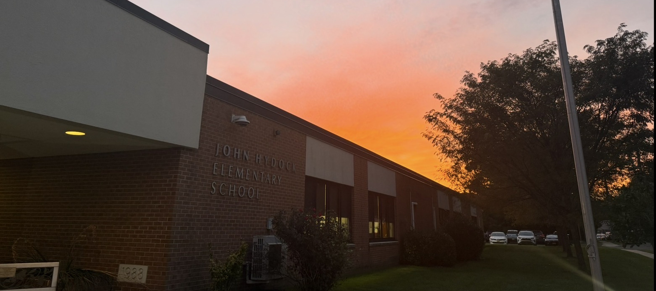 A brick elementary school building stands in the foreground under a vibrant orange and pink sunset sky with a large tree to the right.