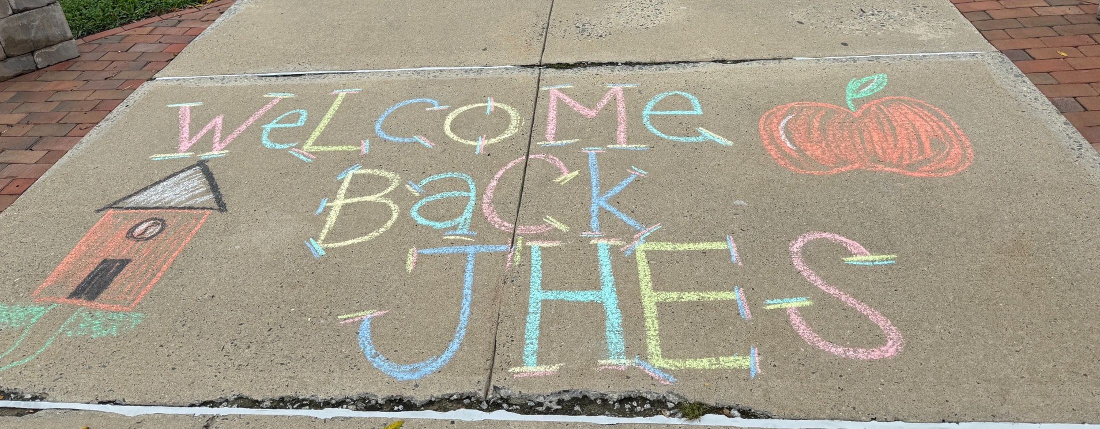 Colorful chalk writing saying "Welcome Back JHES" with a schoolhouse and an apple is drawn on a concrete sidewalk, bordered by red bricks and green grass.