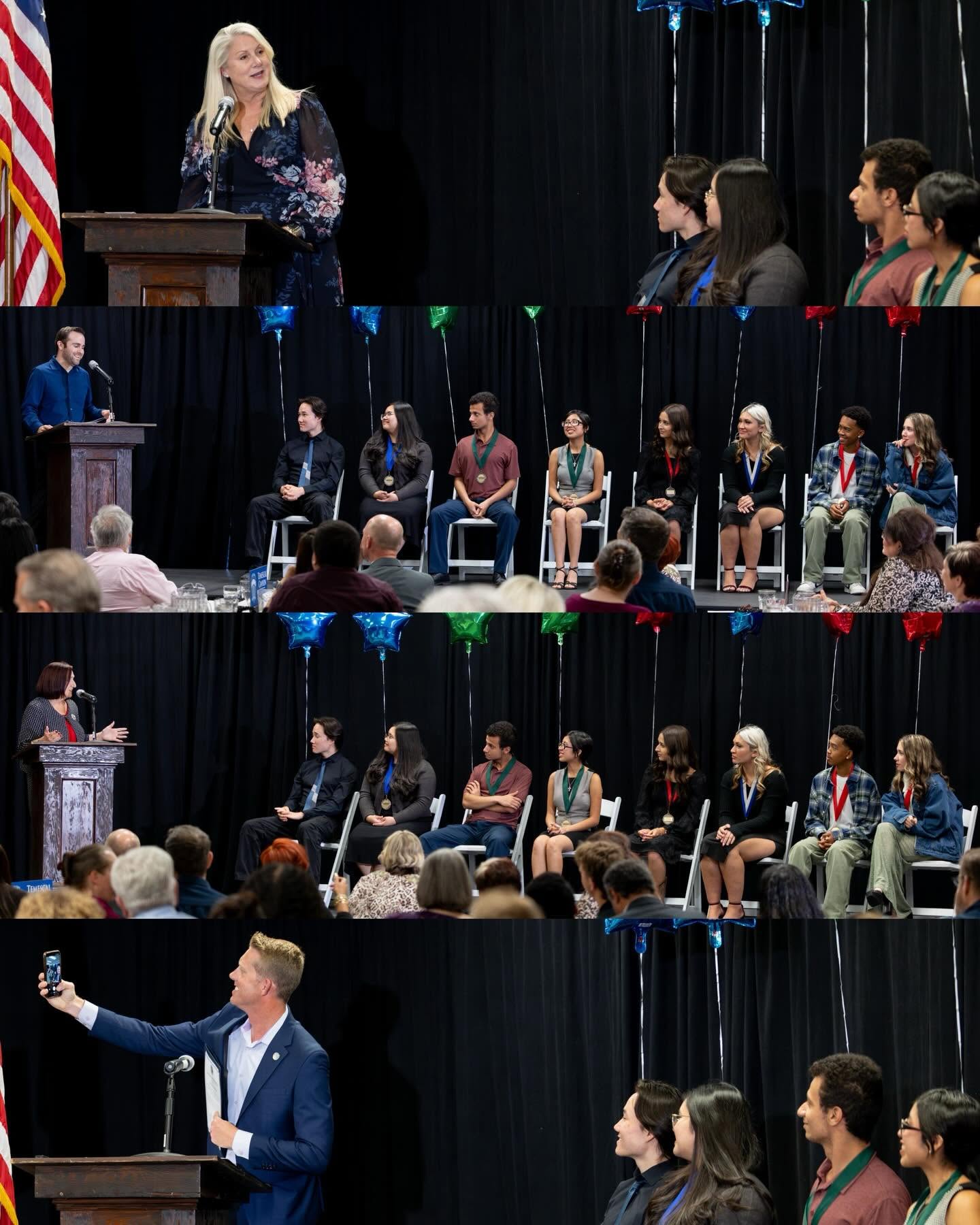 A woman speaks at a podium in the foreground while a group of students sits in chairs against a black curtain background.