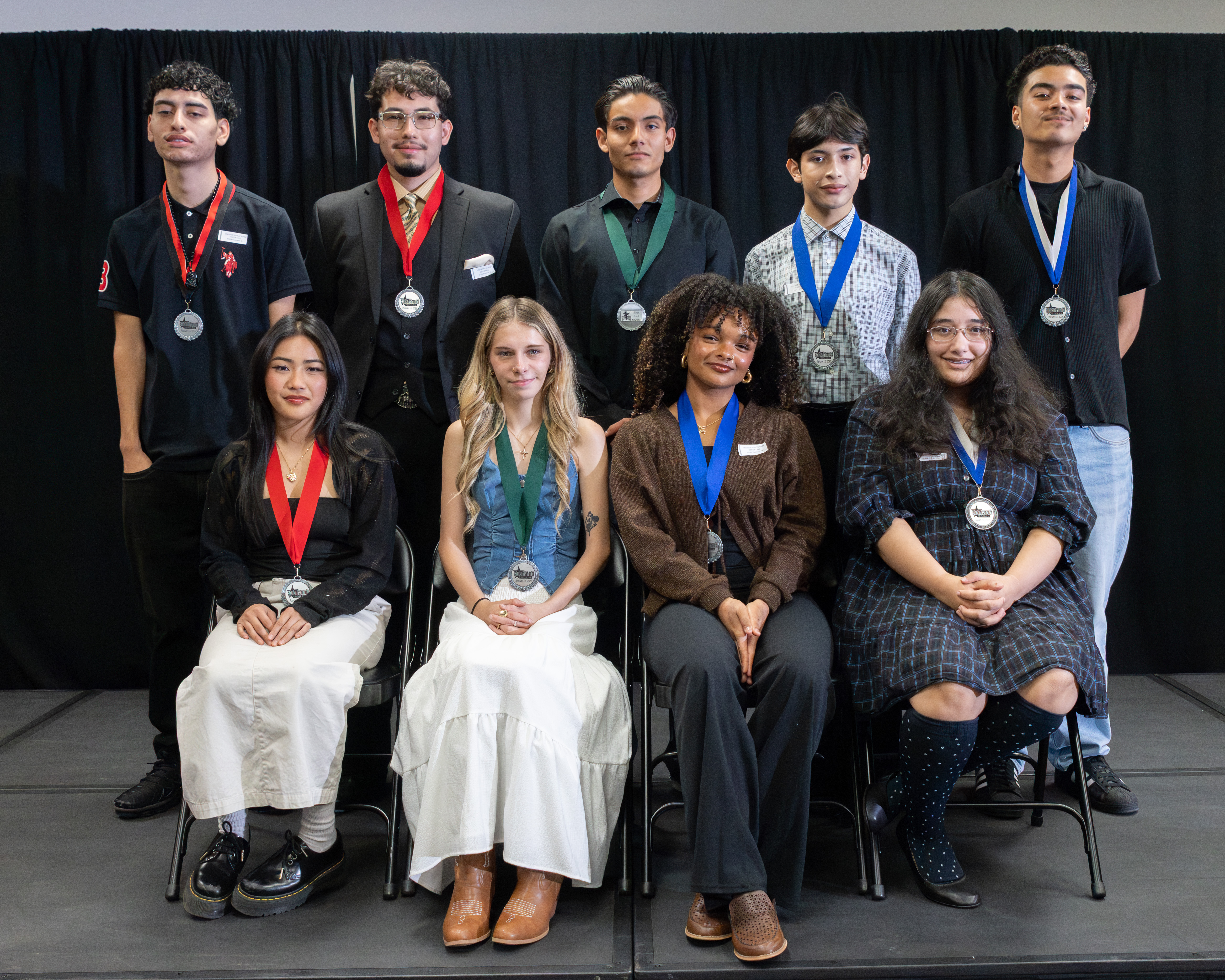 A group of young adults wearing medals pose for a photo against a black curtain backdrop.