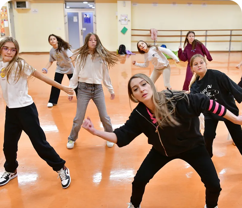 A group of young women in casual athletic wear are dancing in a spacious studio with an orange floor and pale walls.
