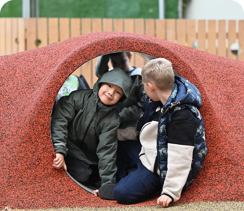 Two young boys play inside a red textured tunnel, with a wooden fence and green foliage in the background.