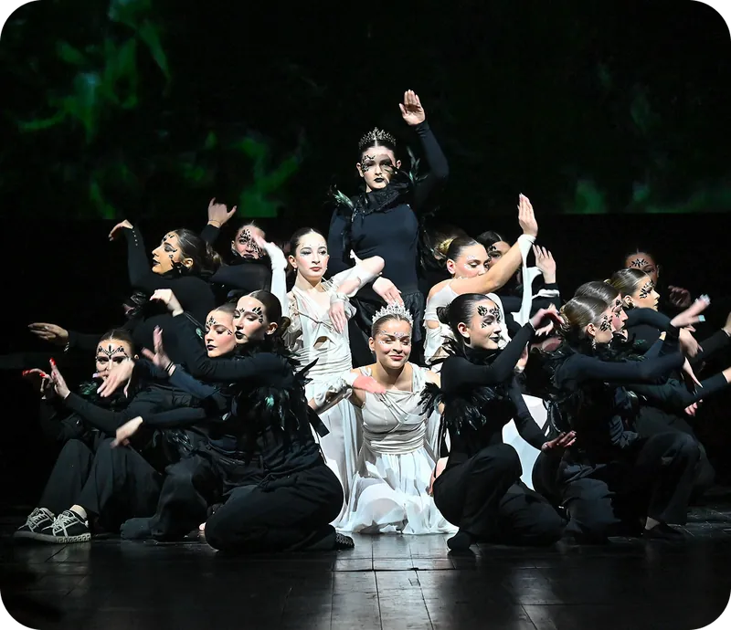 A group of dancers in black and white costumes perform on a dark stage against a green-tinted background.