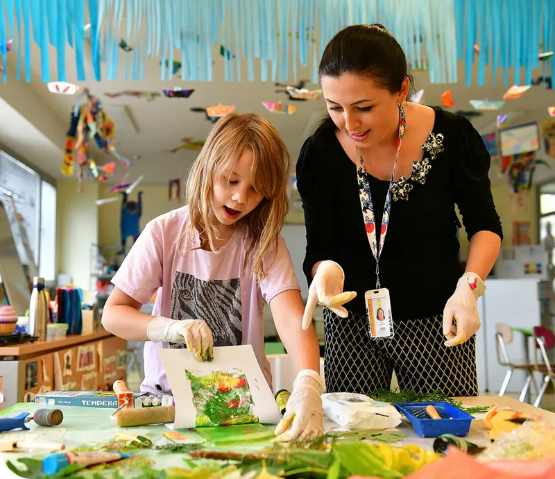 A young girl and a woman wearing gloves work on an art project in a classroom decorated with blue streamers.