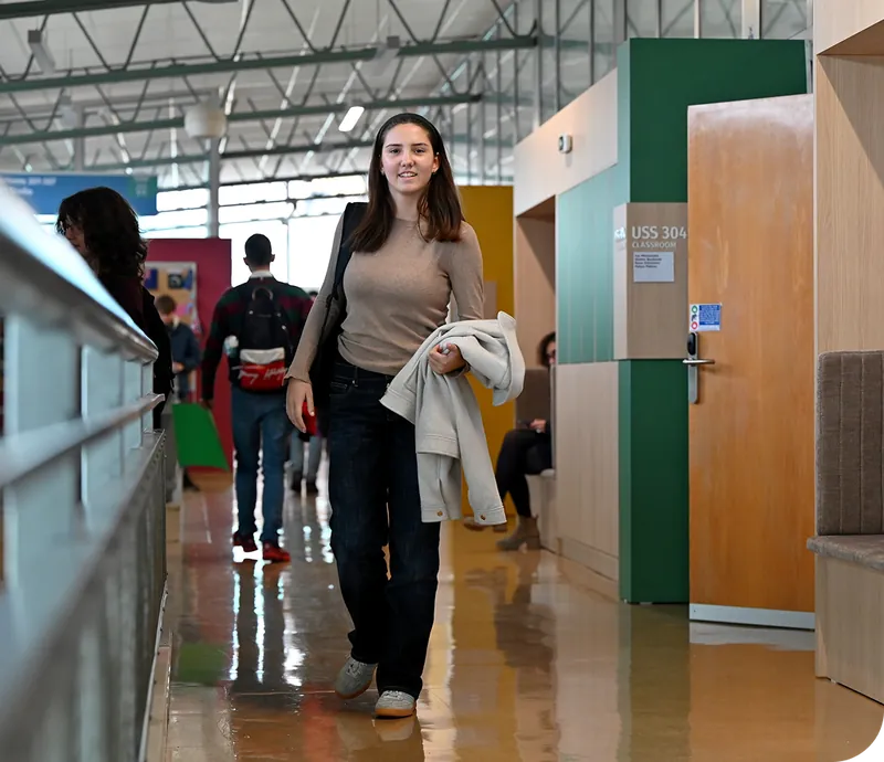A woman in a tan shirt and dark pants walks through a modern hallway with other students and classroom doors in the background.