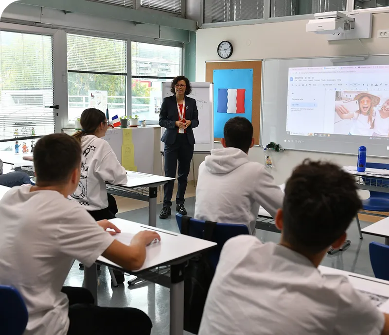 Students in white shirts sit at desks in the foreground, facing a teacher standing before a whiteboard and a French flag in the background.