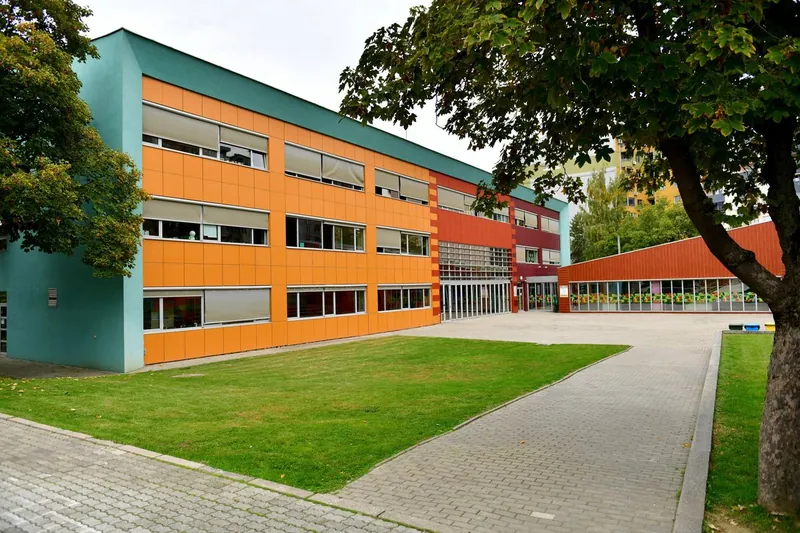 A green lawn and paved walkway lead toward a modern, multi-colored school building framed by leafy green trees.
