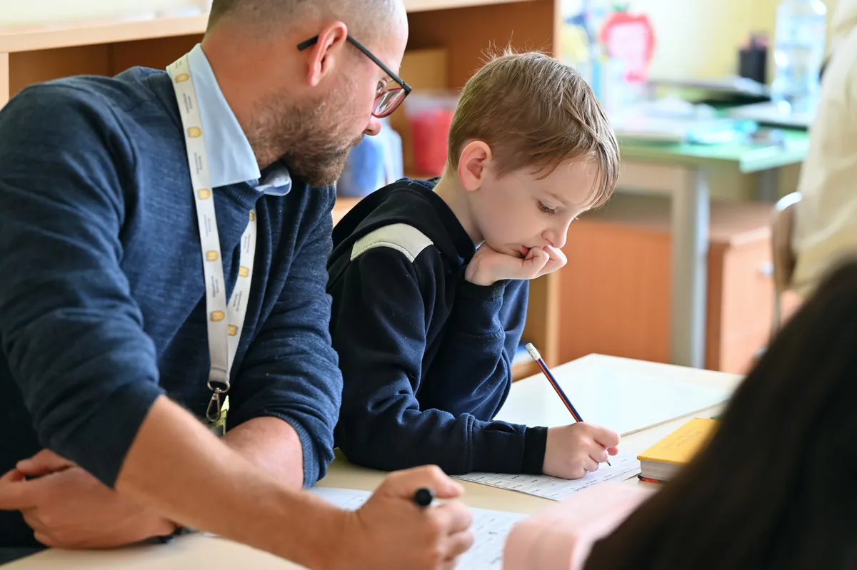 A man and a young boy work together at a table in a brightly lit classroom.
