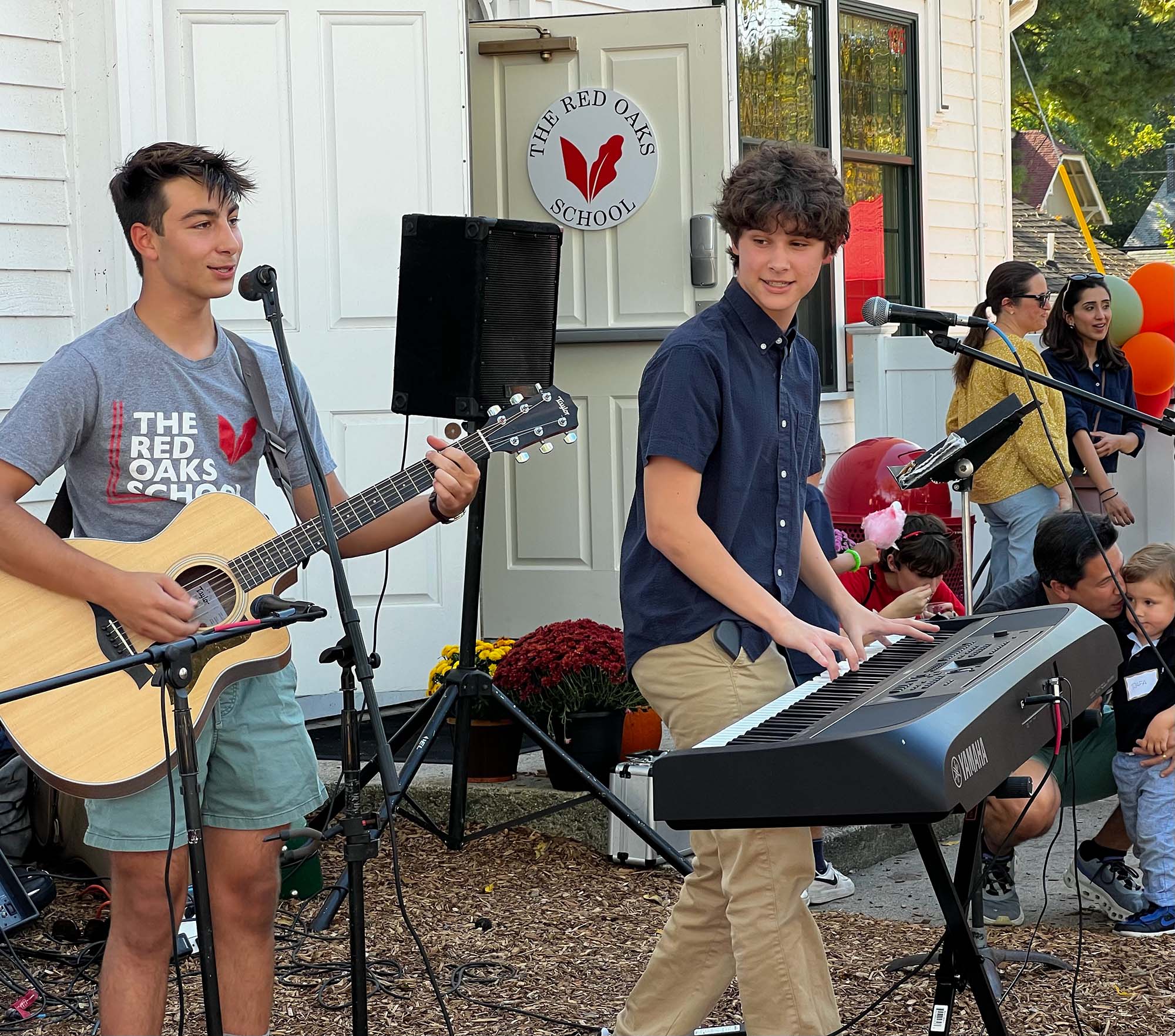 Two young men perform music with a guitar and keyboard in the foreground, while a crowd and a building with "The Red Oaks School" logo stand in the background.