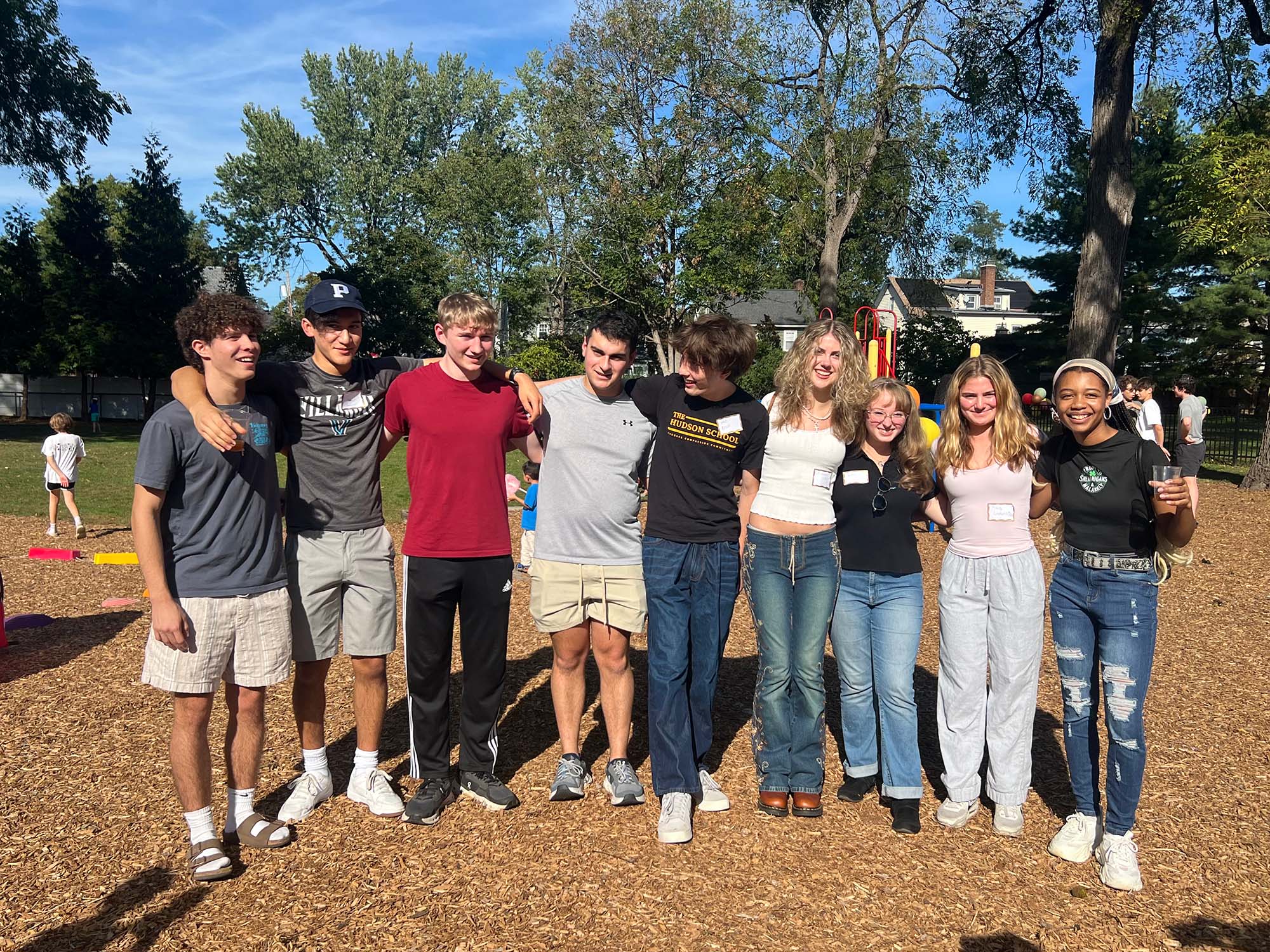 A group of smiling teenagers stands together on a wood-chipped ground with green trees and a house in the background.