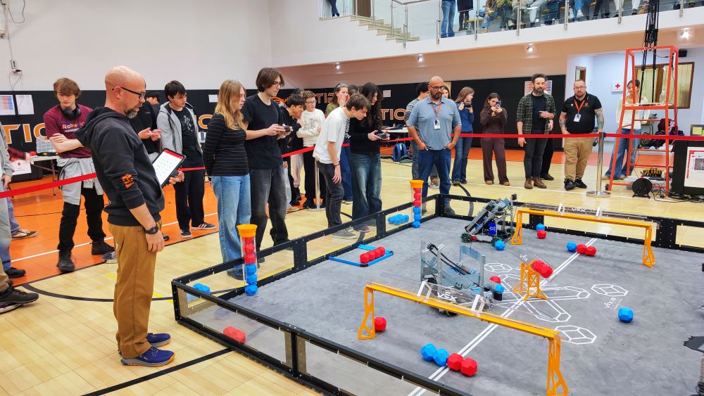 A group of people watch a small robot compete on a marked game field inside a brightly lit gymnasium.