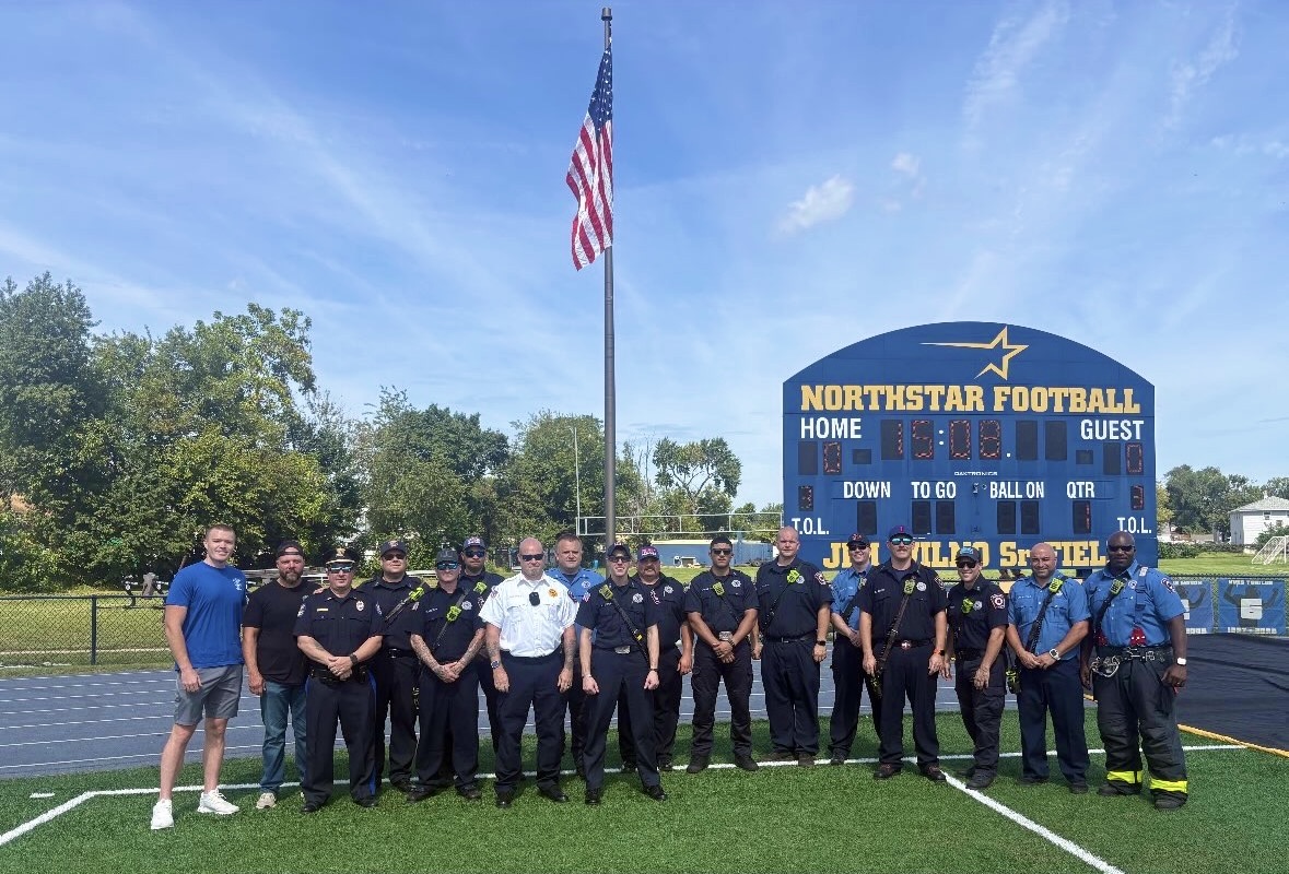 A group of uniformed officers and a man in a blue shirt stand on a football field in front of a large scoreboard and an American flag.