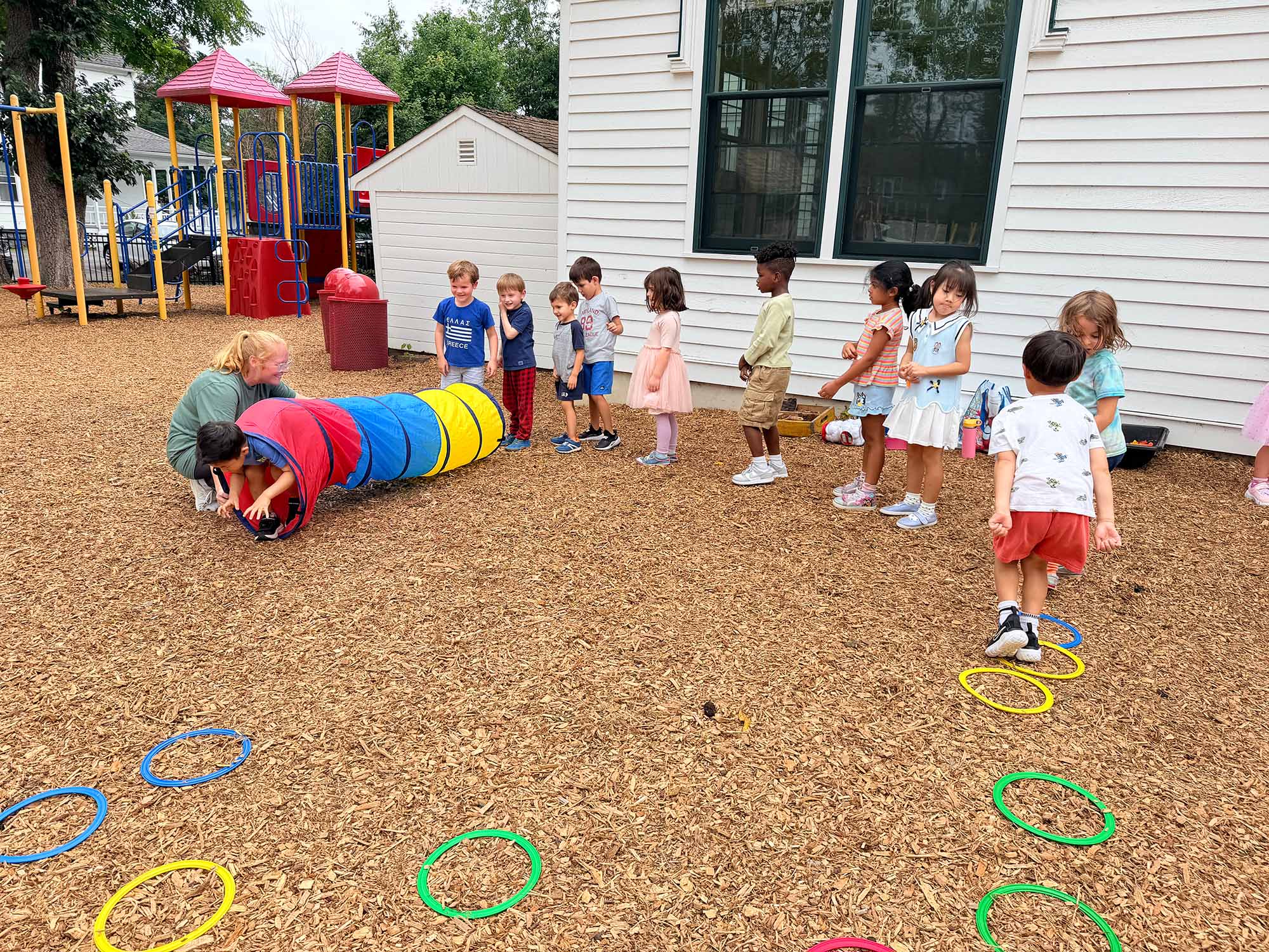 Children play with a colorful tunnel and hula hoops on a woodchip playground in front of a white house and play structure.